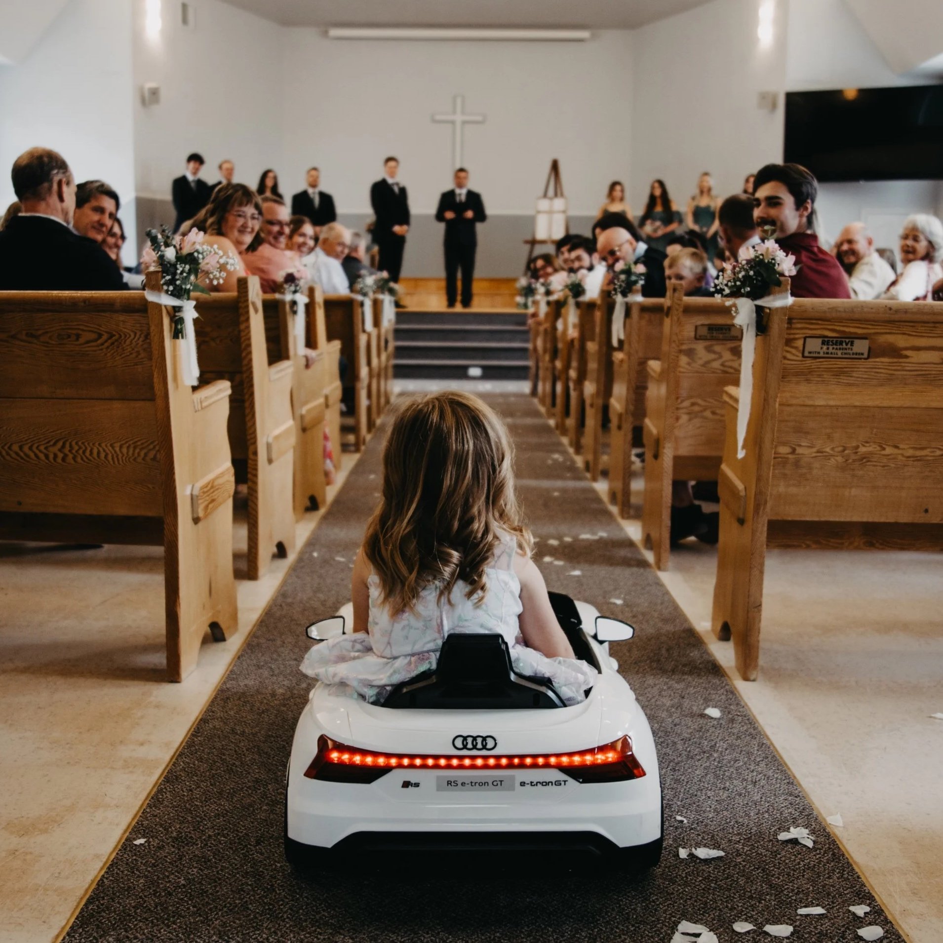 A young girl sitting in a toy car on a church aisle, facing an altar with a cross, attending a wedding ceremony with guests smiling and watching.