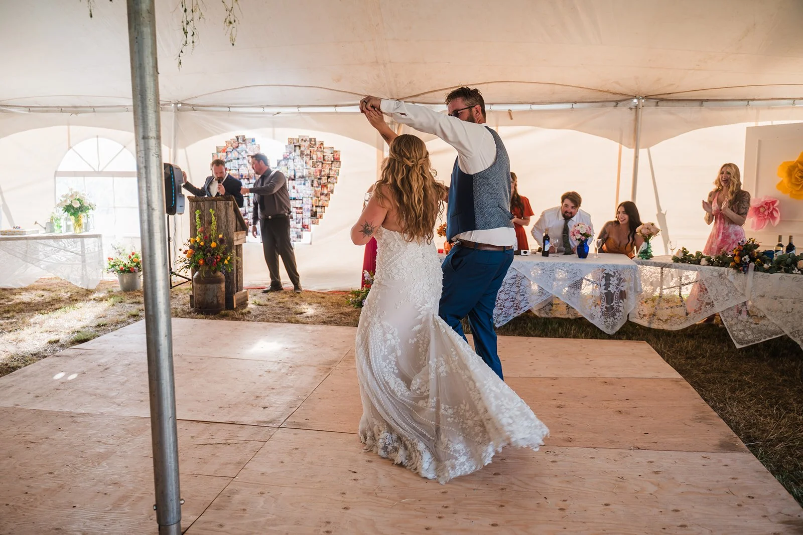 A bride and groom dancing on a wooden dance floor at their wedding reception inside a decorated tent with a head table and guests watching.
