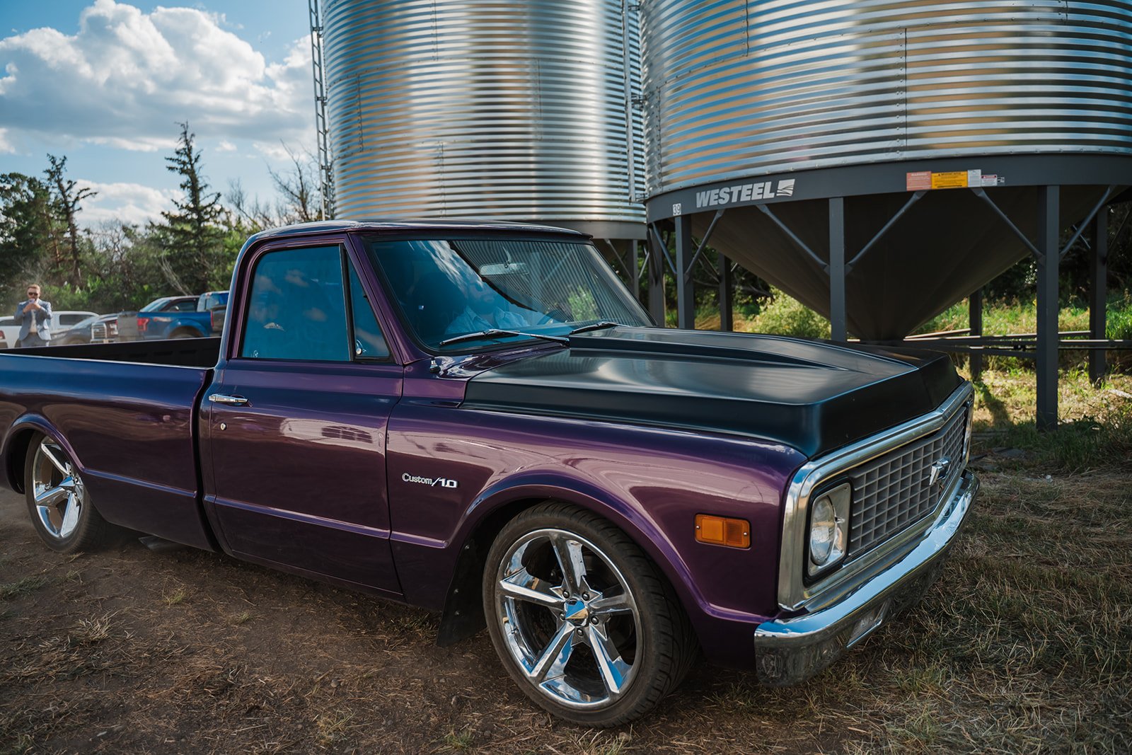 A vintage purple and black Chevrolet pickup truck parked on dirt with large silver grain silos in the background.