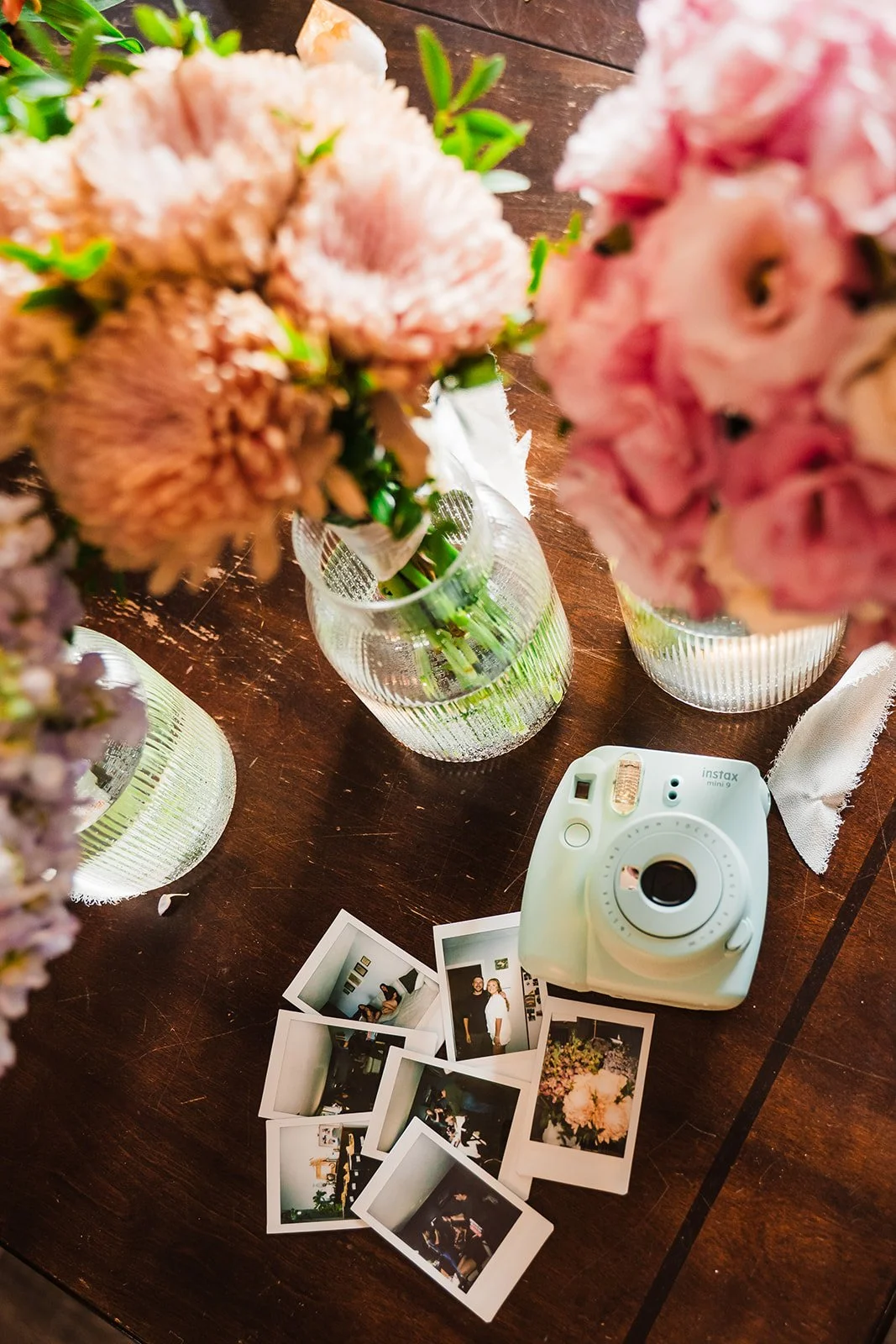 Clothes of pink color with white flowers, a white instant camera, several polaroid photos, and vases of pink and purple flowers on a dark wooden table.