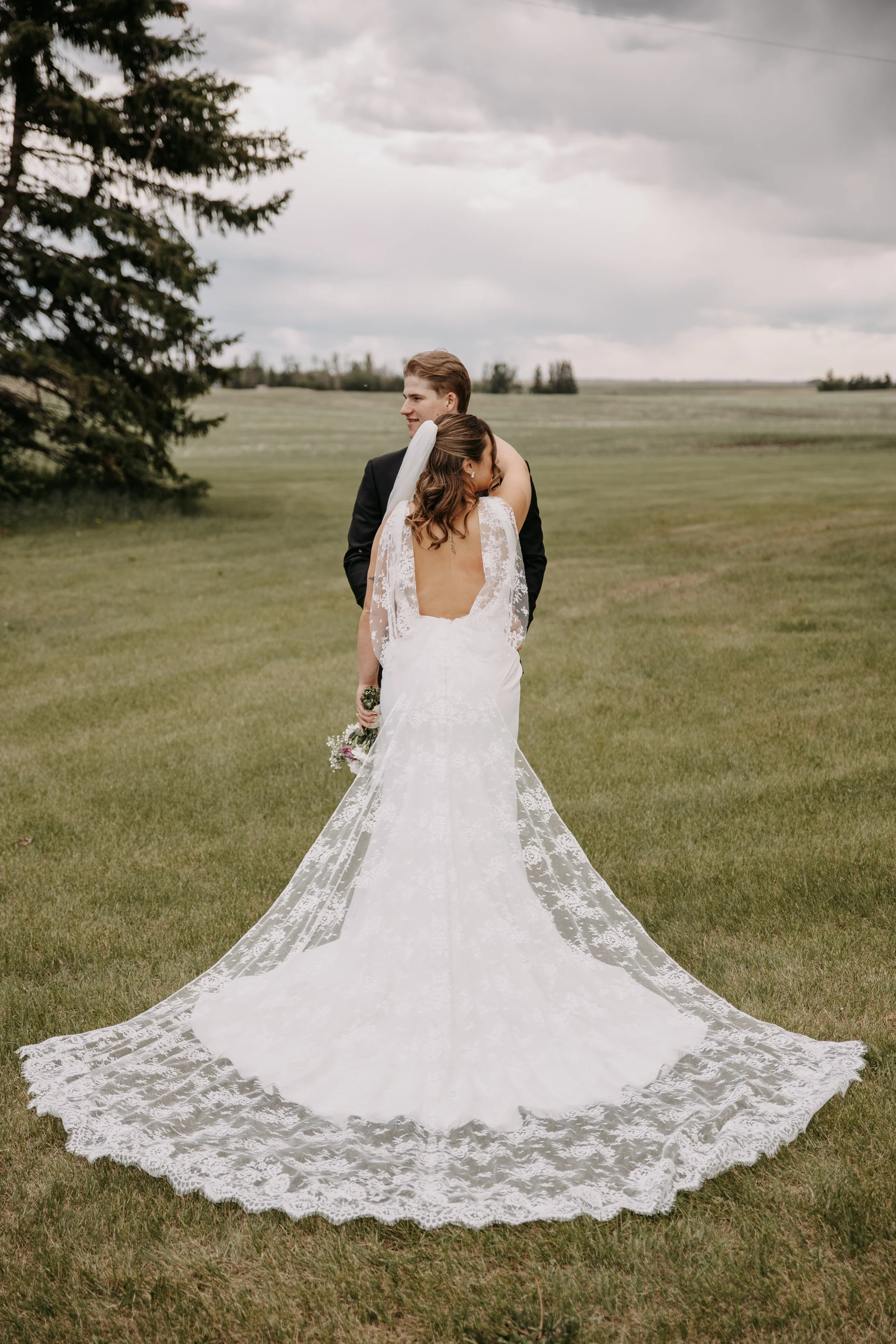A bride in a white lace wedding gown and a groom in a dark suit embracing outdoors on a grassy field with overcast skies.