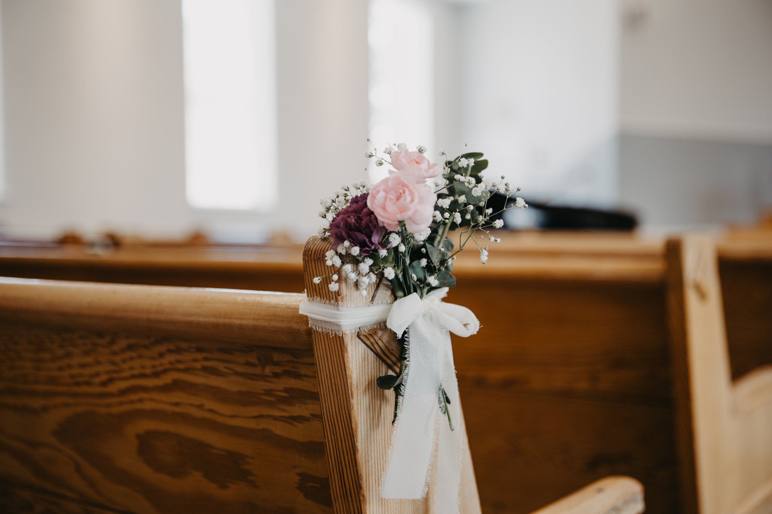 A wooden church pew decorated with a small bouquet of pink and purple flowers, white baby's breath, and green leaves, tied with a white ribbon.