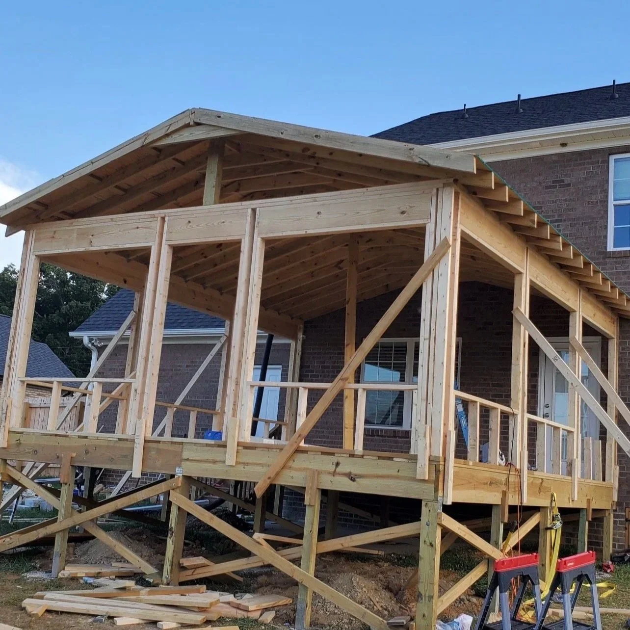 A wooden deck under construction attached to a brick house, with support beams, railing, and a roof structure in place.