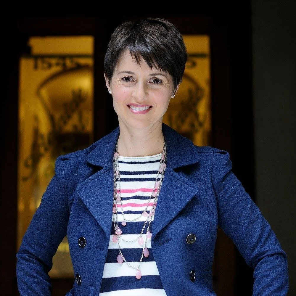 A woman with short dark hair smiling, wearing a navy blue blazer over a striped shirt and layered necklaces, standing indoors with a blurred warm-toned background.
