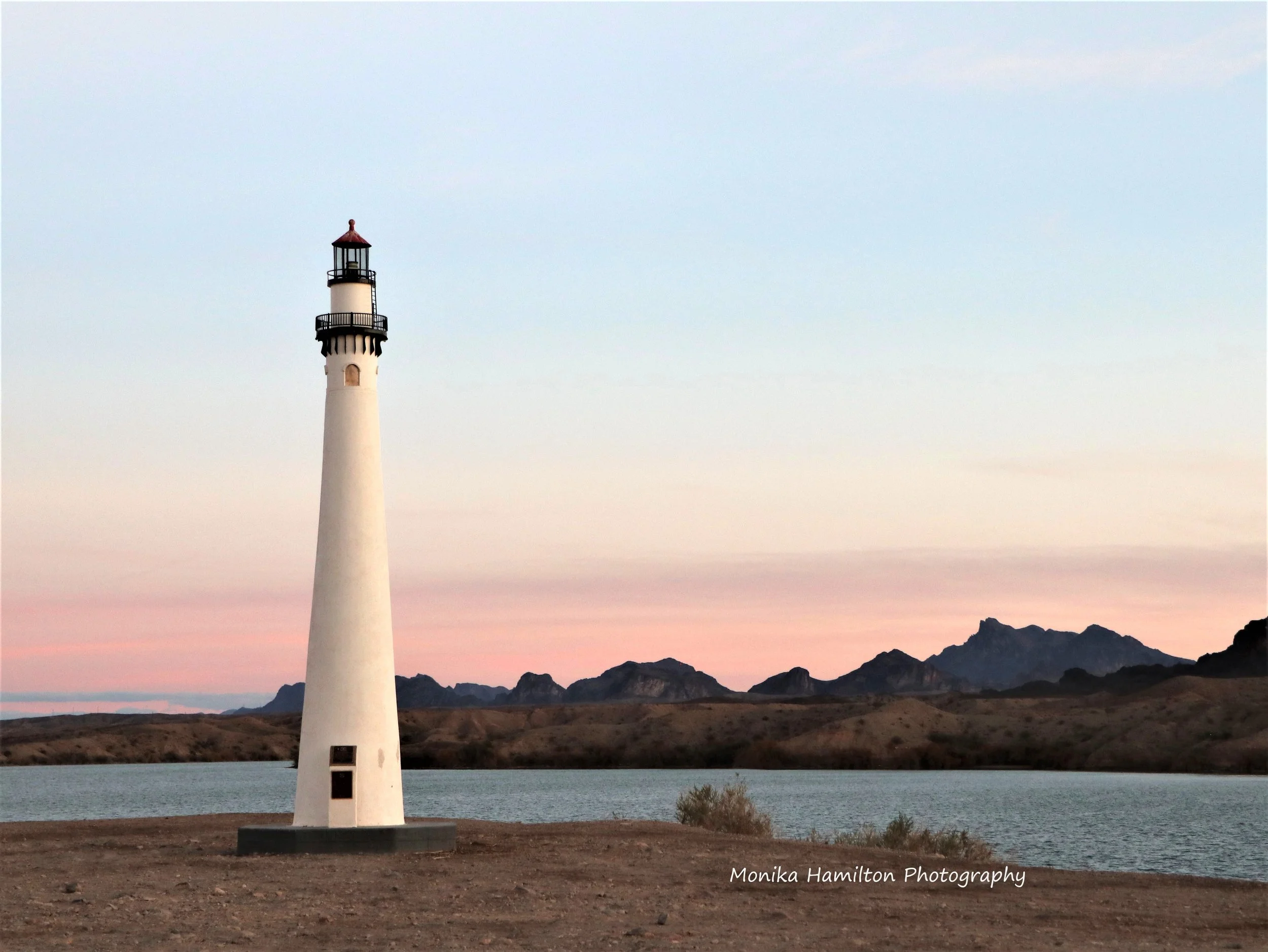 A tall white lighthouse standing on Lake Havasu, Arizona's shore with mountains in the background, and a pastel pink and blue sky at dusk. Monika Hamilton Photography