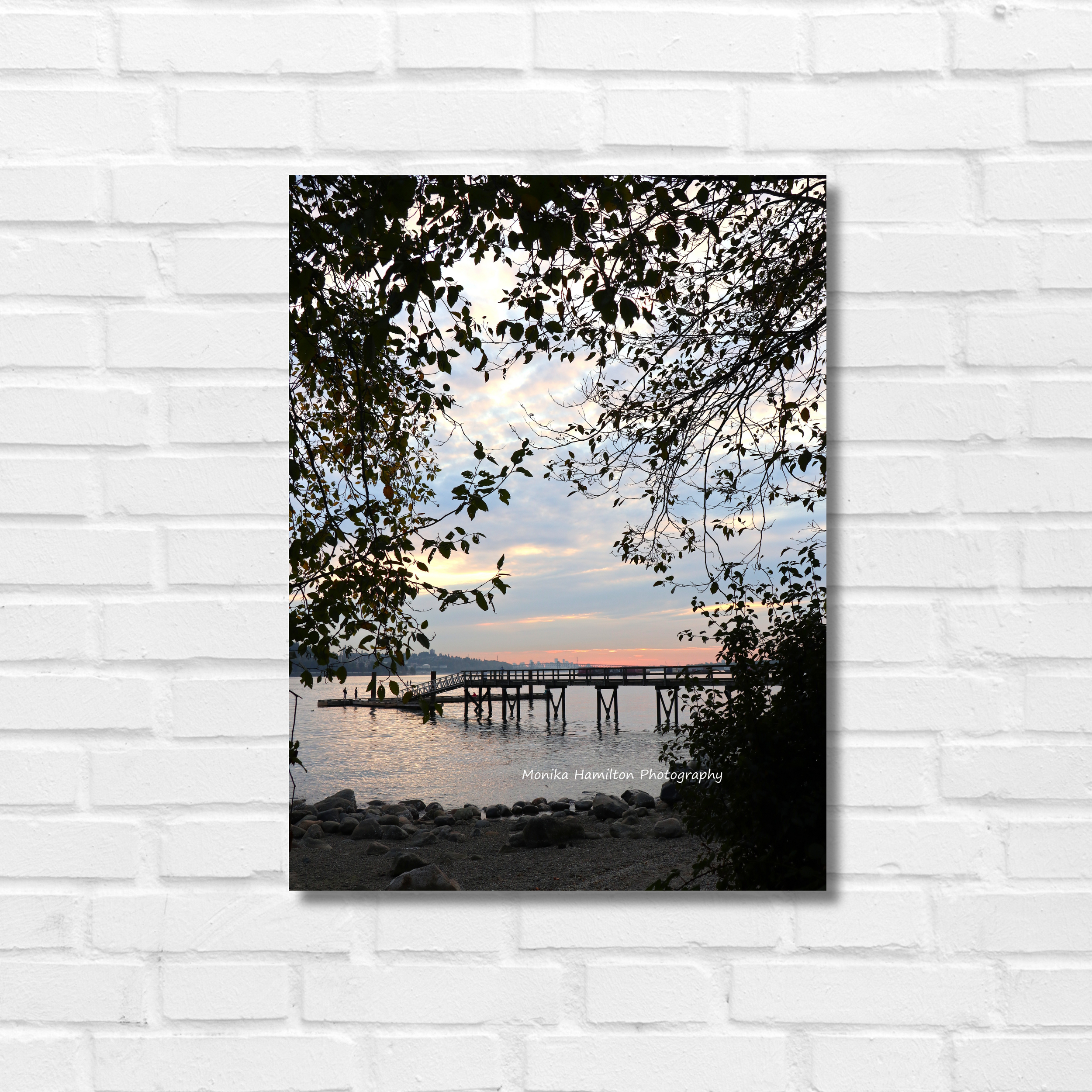 Sunset at Cate's Park in North Vancouver, British Columbia. The pier is framed by overhanging branches and leaves, with a partly cloudy sky, pastel sunset, and city skyline in the background. Monika Hamilton Photography