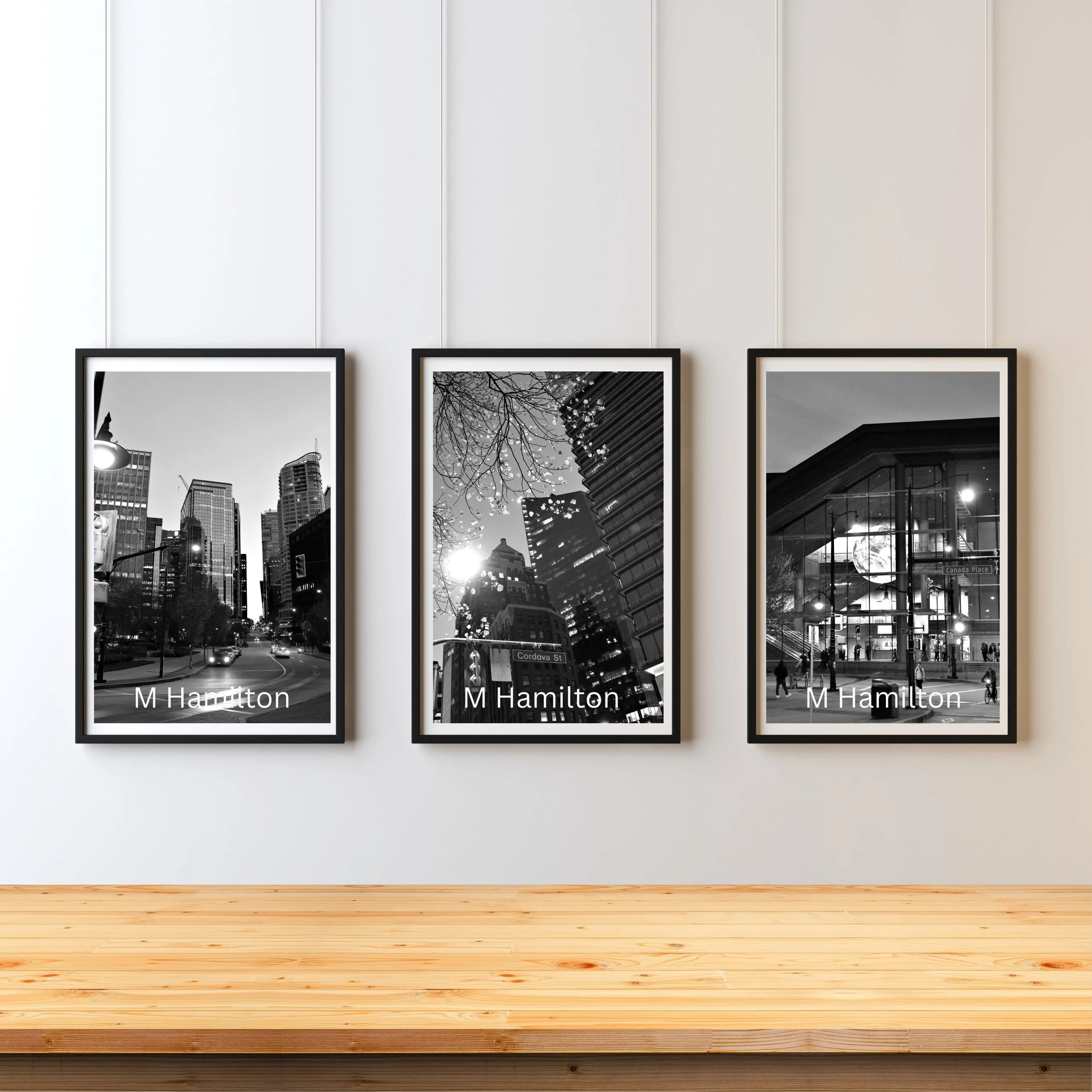 Three black and white framed cityscape photographs on a white gallery wall, with a wooden floor below.