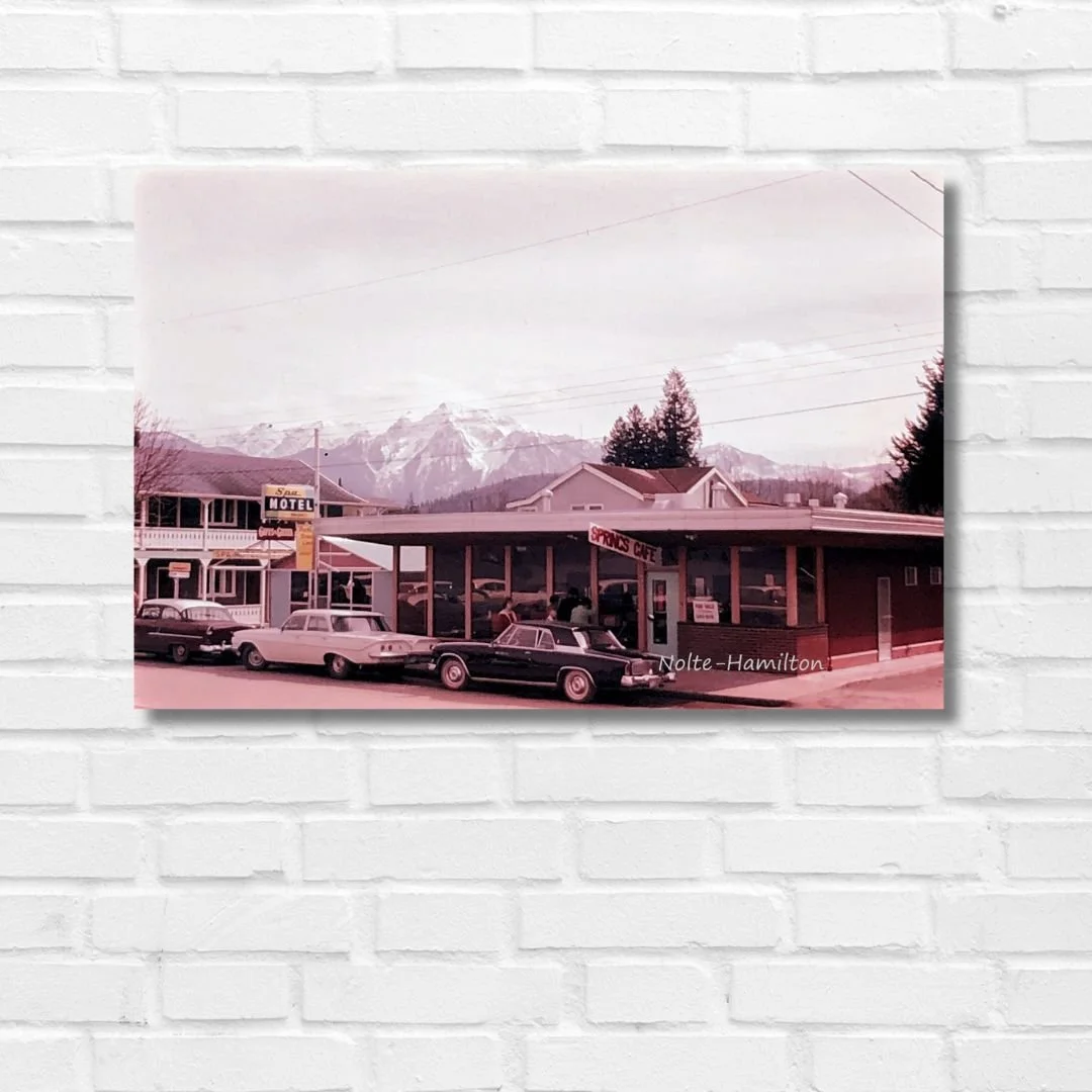 A vintage photo of the Springs Cafe in Harrison Hotsprings. This original city scene was taken by my dad in the 1960s. There are mountains in the background, classic cars parked in front of the cafe, and signs for a motel and a café.