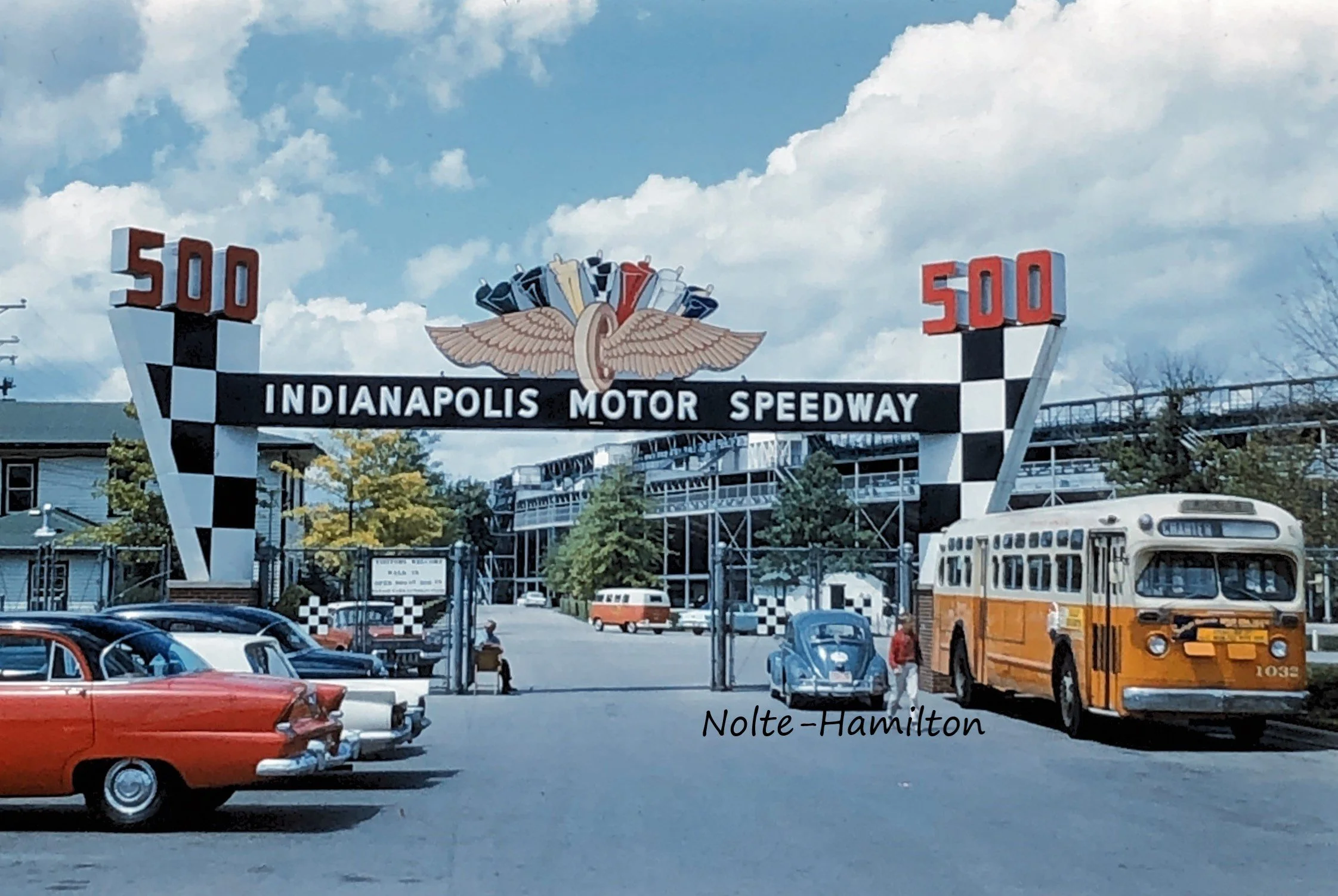 Vintage 1960s entrance sign for Indianapolis Motor Speedway with checkered flag design, vintage cars parked below, and a yellow bus, under a partly cloudy blue sky. Original Indy 500 photography. Monika Hamilton Photography