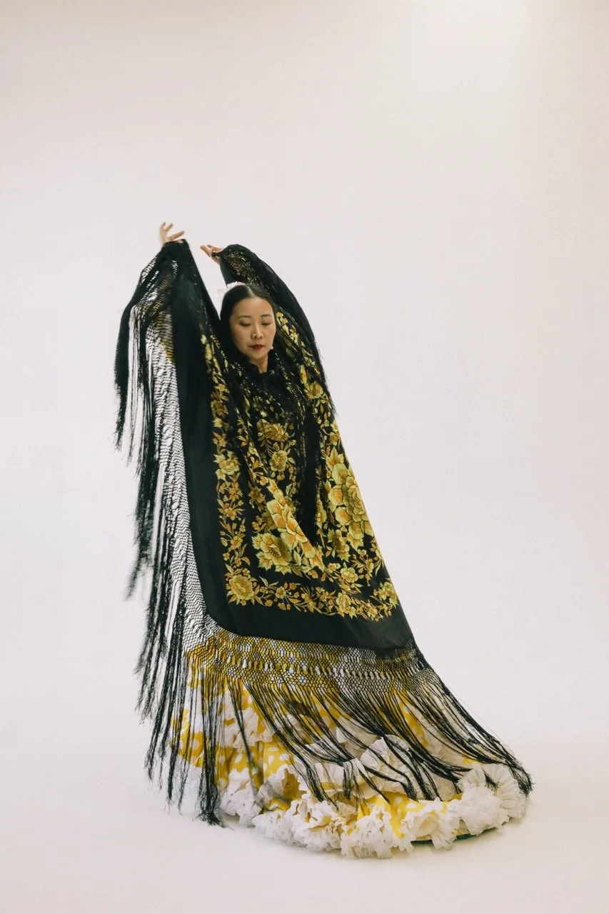 A woman dressed in an elaborate black and gold embroidered dress with a long, fringed black shawl, posing with her eyes closed and arms raised against a plain white background.