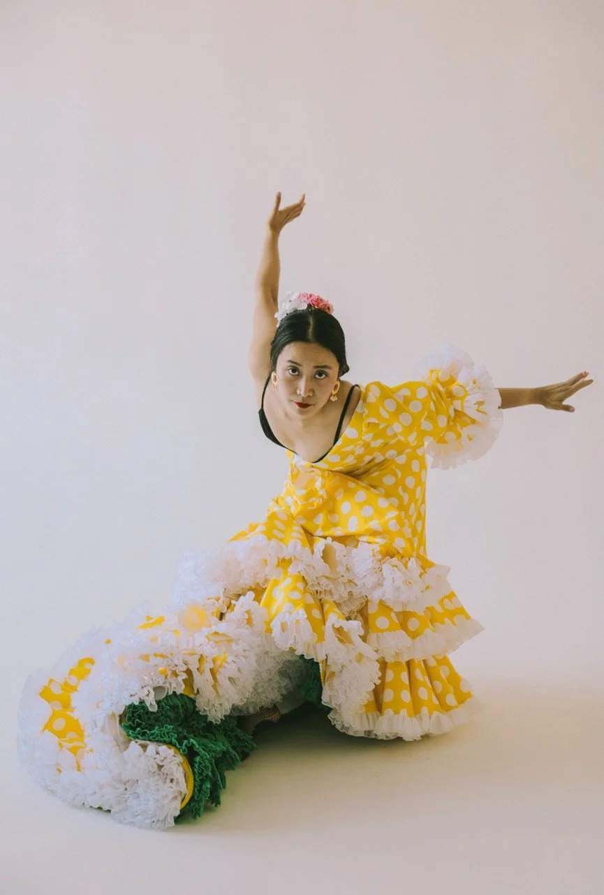 A woman in a yellow polka dot dress with ruffled white accents performs a dance pose against a plain white background.