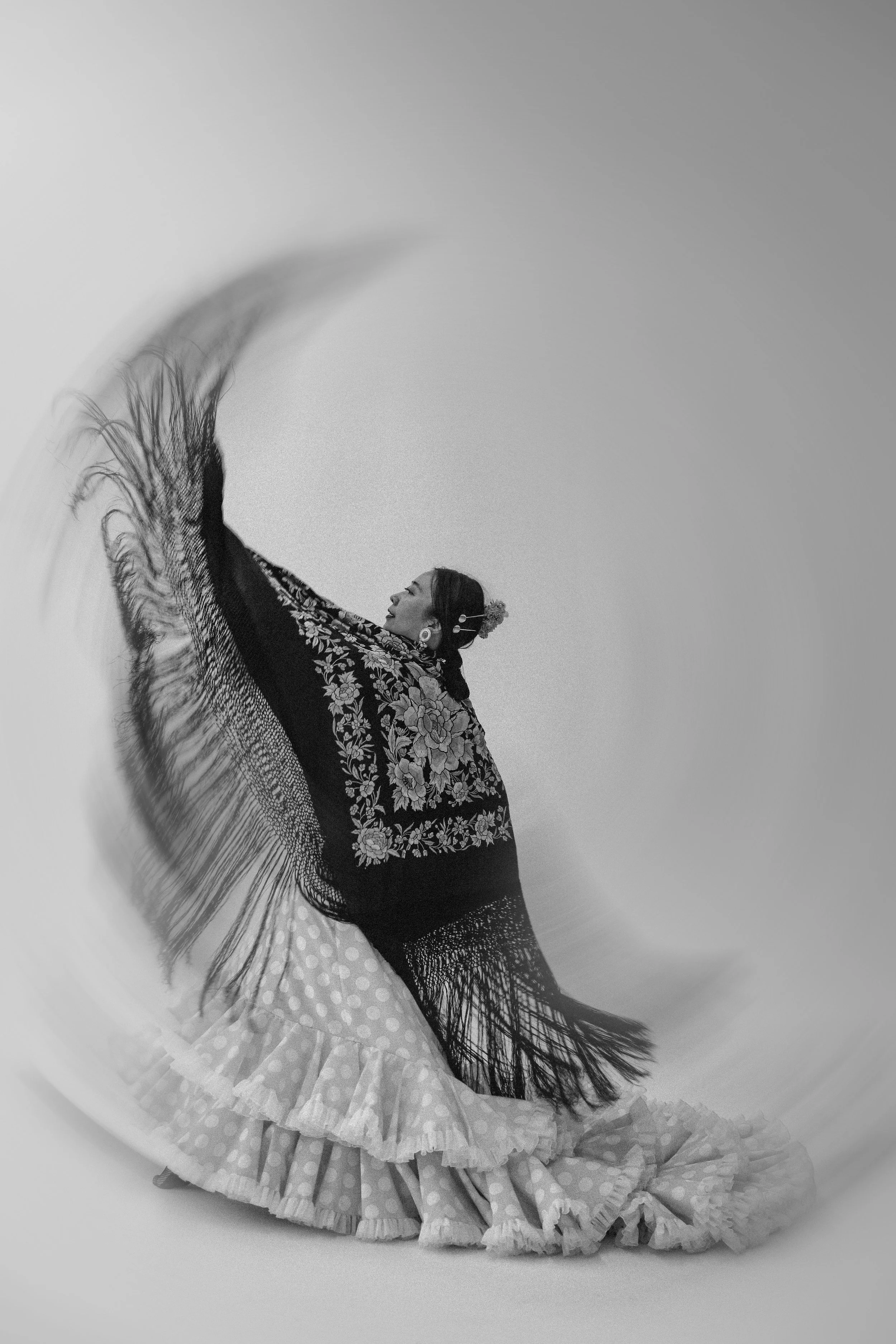 A woman in traditional Mexican dress spinning with a large shawl, creating a circular motion blur in black and white.