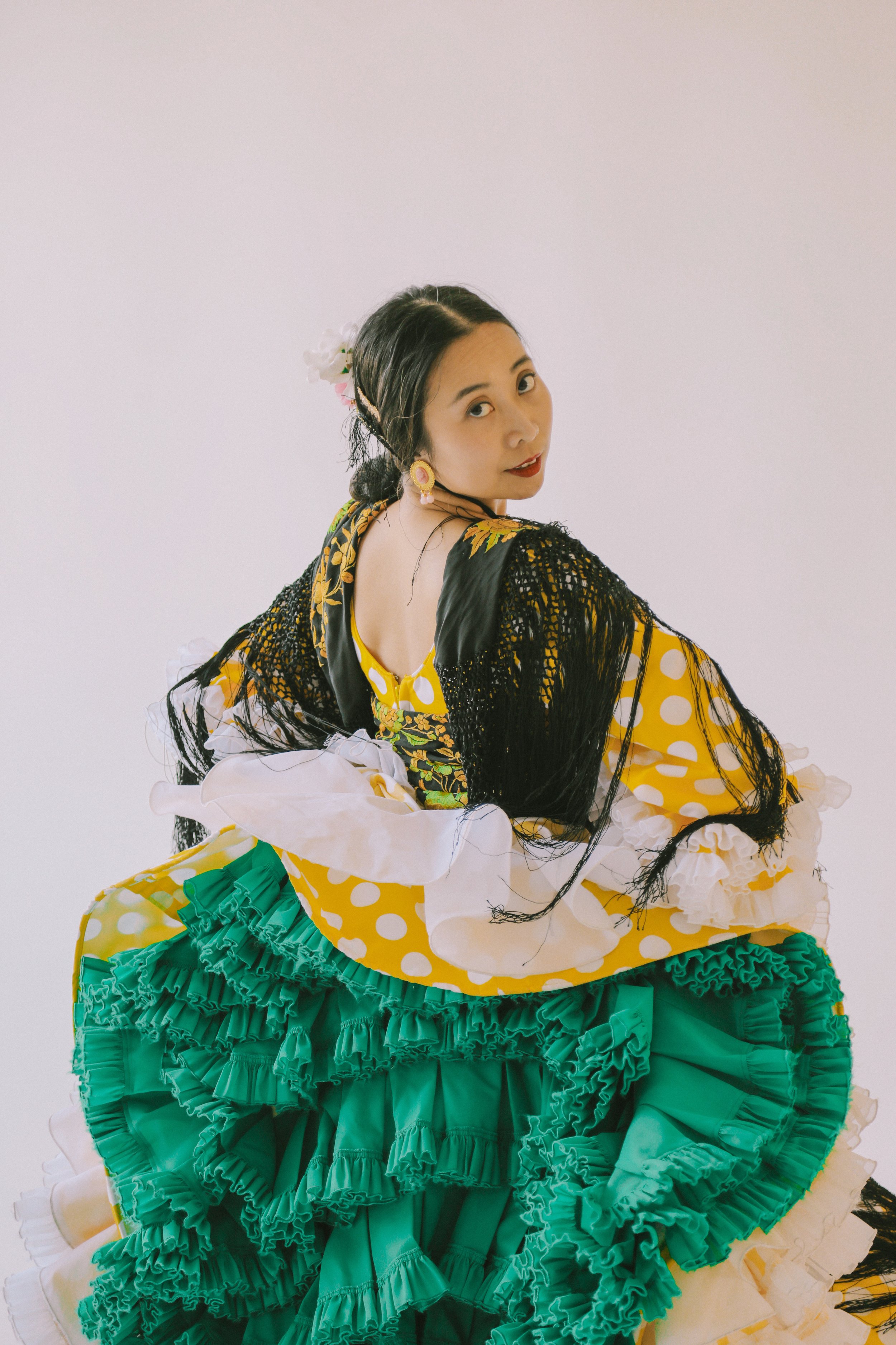 Woman in traditional Mexican dress with yellow polka dots and green ruffles, posing against a plain white background.