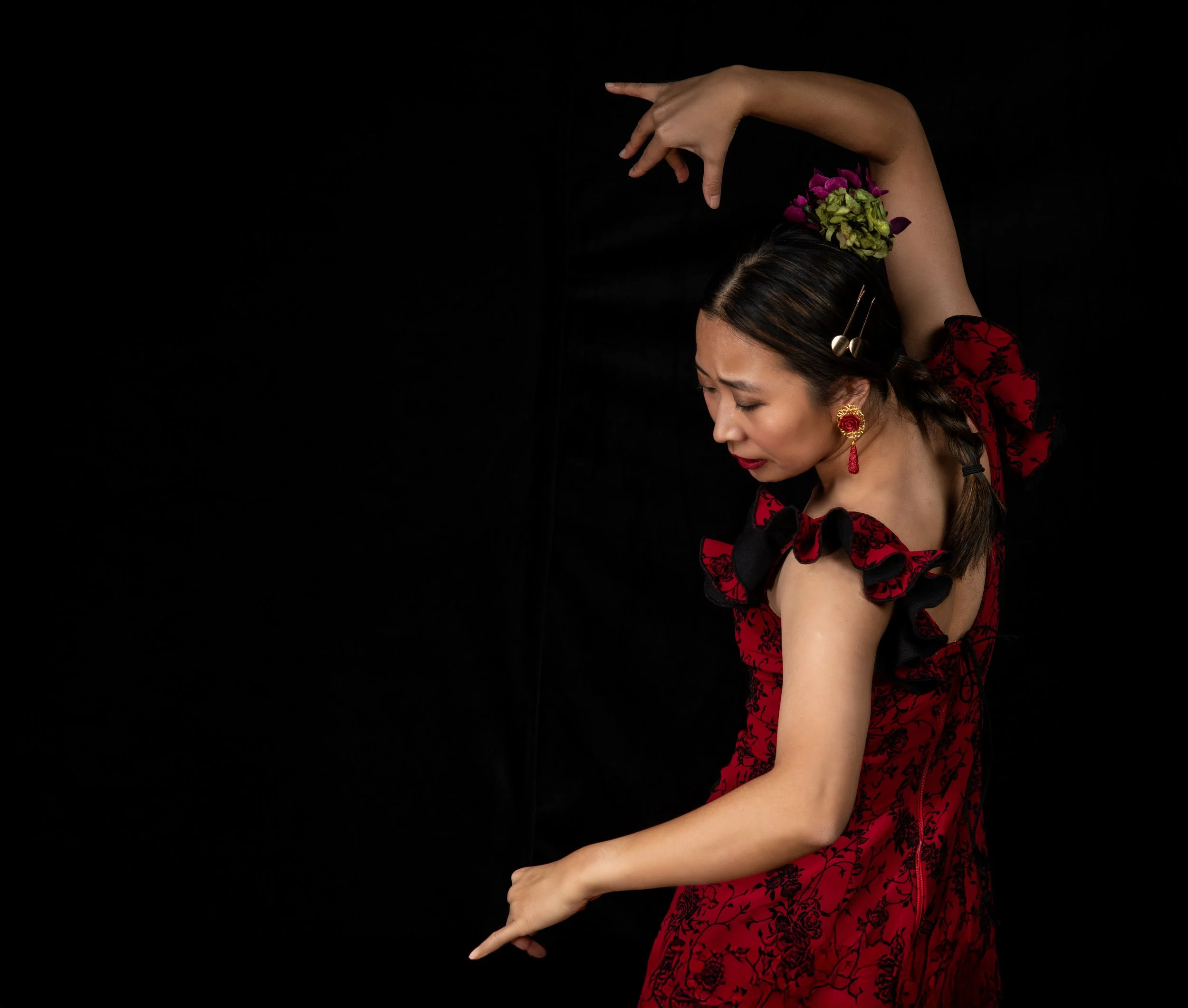 A woman in a red and black dress with floral patterns, jewelry, and flowers in her hair, poses against a black background with her eyes closed and arms raised in a dance or expressive pose.