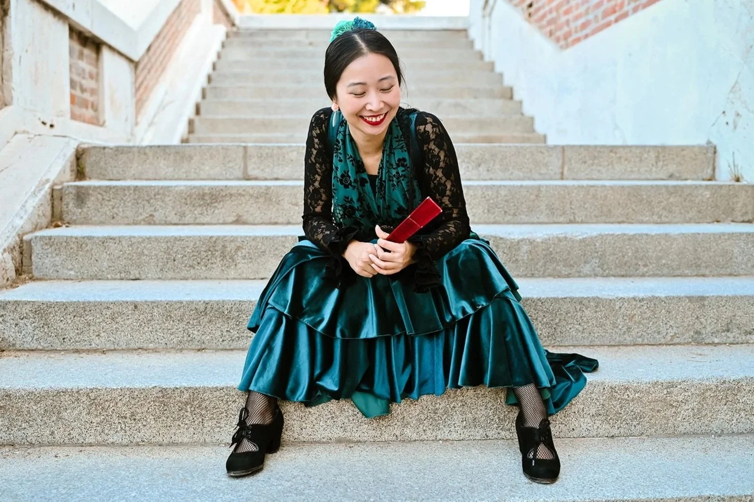 A woman in a teal satin dress with black lace top and black shoes sitting on outdoor concrete stairs, smiling while looking at her phone.