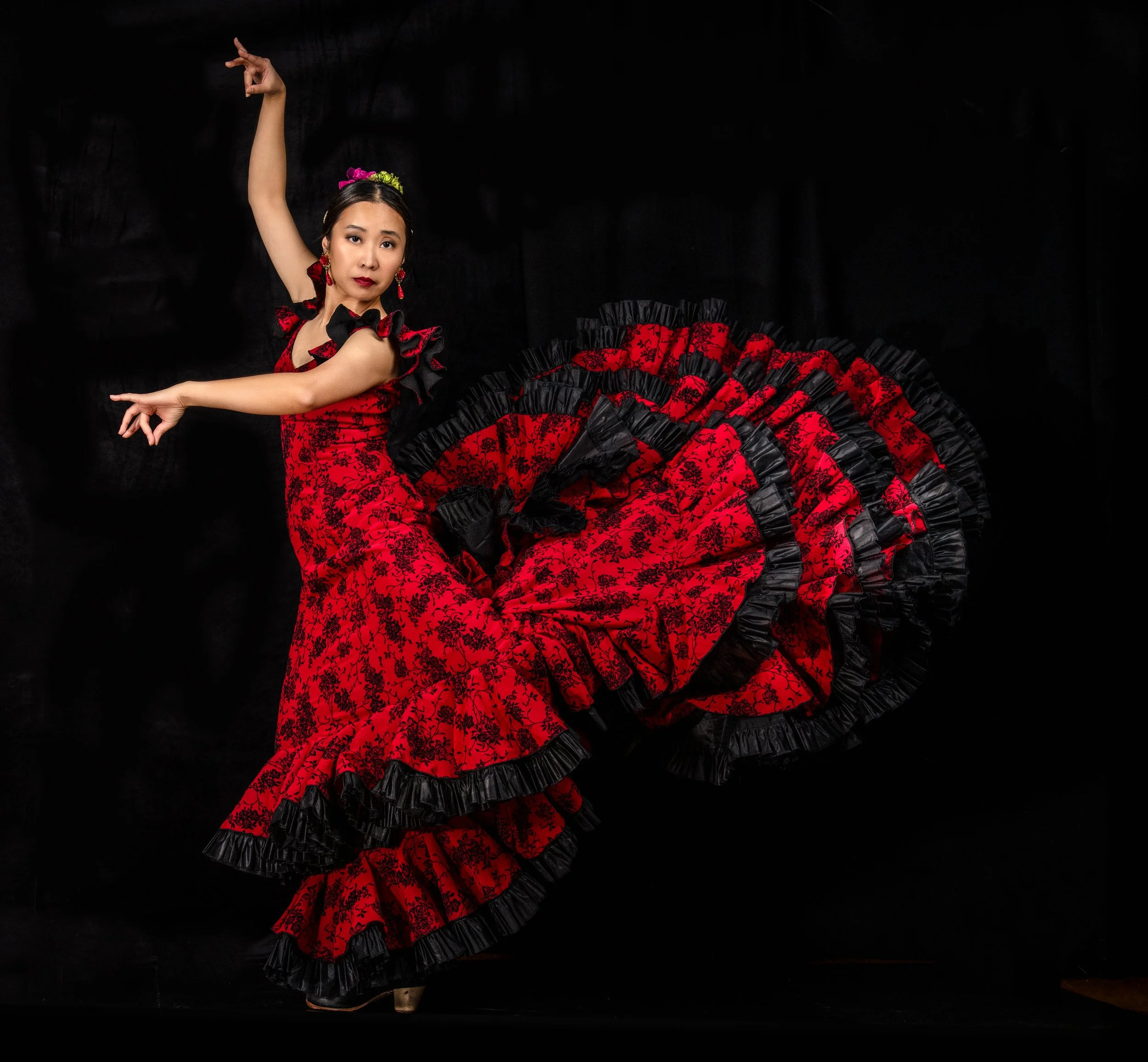 A woman performs a traditional Mexican folkloric dance, wearing a red and black ruffled dress with intricate patterns, holding her skirt in an elegant pose against a black background.