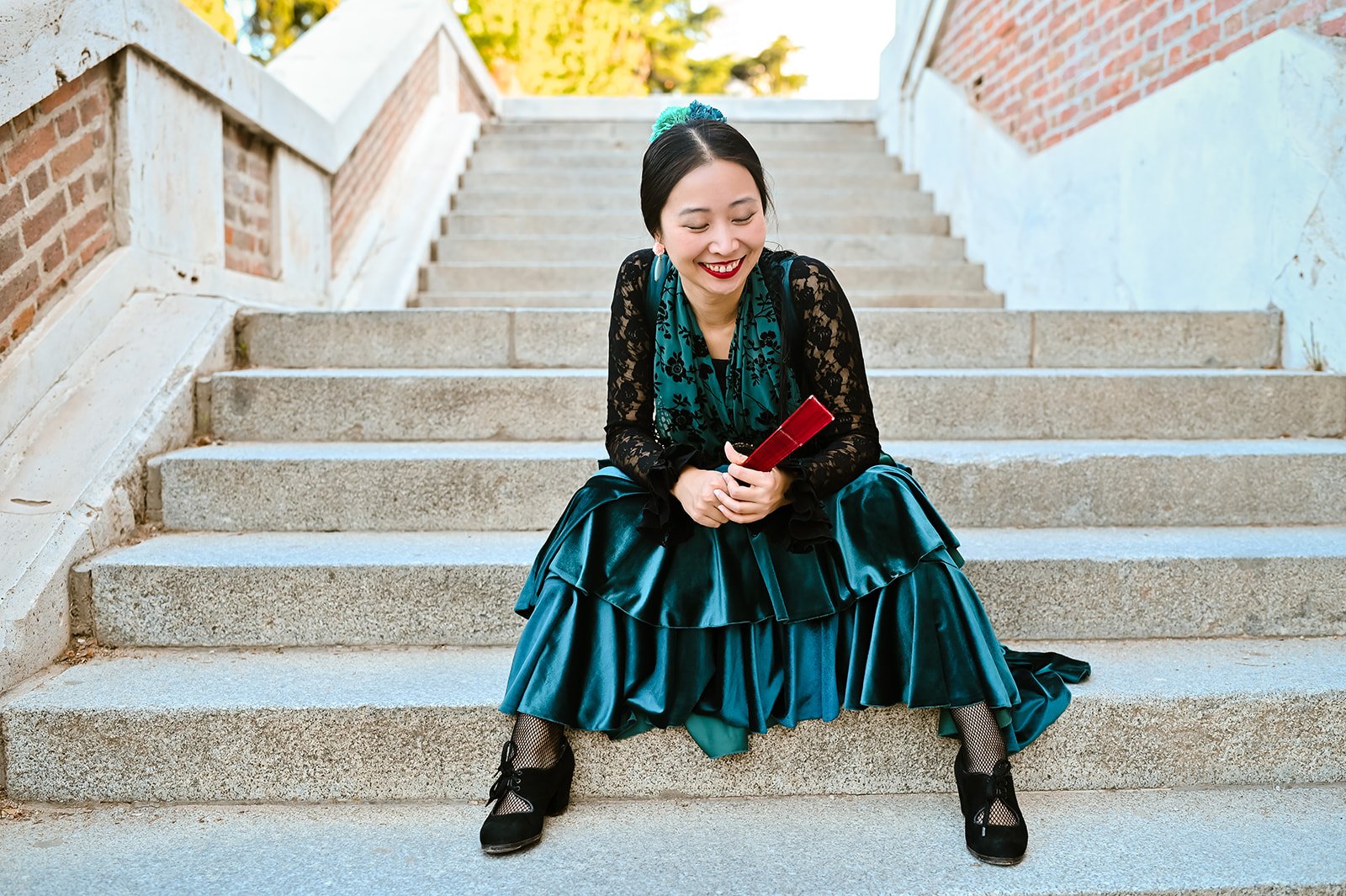 A woman sitting on outdoor concrete stairs, smiling and holding a red book. She is wearing a teal dress with a matching teal flower hair accessory, black lace top, black shoes with bows, and fishnet stockings.