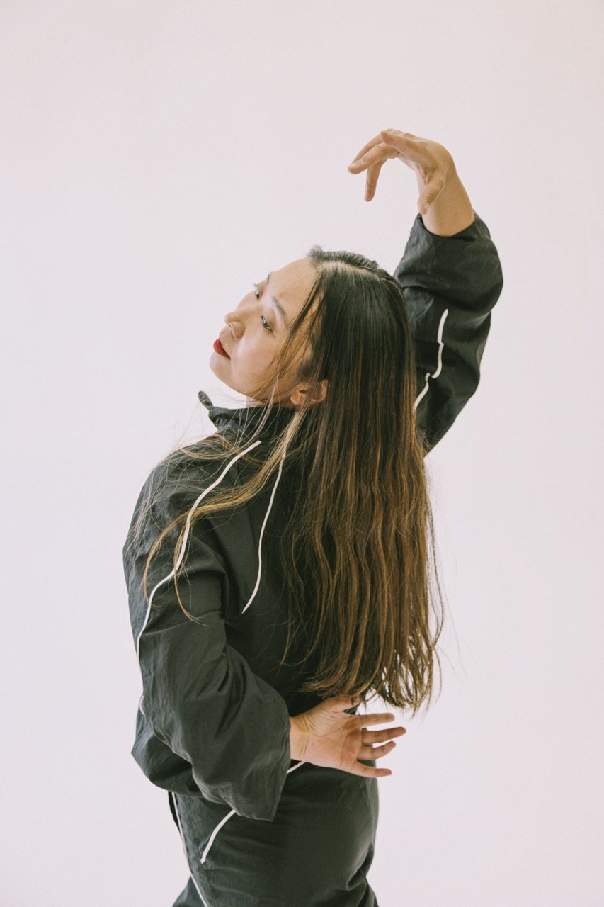A woman with long brown hair wearing a black athletic jacket and pants, posing with her hand on her hip and her other arm raised above her head against a plain white background.