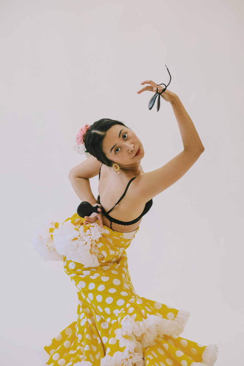 A woman with dark hair in an updo, adorned with pink flowers, wearing a black bikini top and a yellow polka dot skirt with ruffles, poses with her left arm raised, holding a pair of sunglasses, and her right arm bent behind her, holding her skirt. Sh