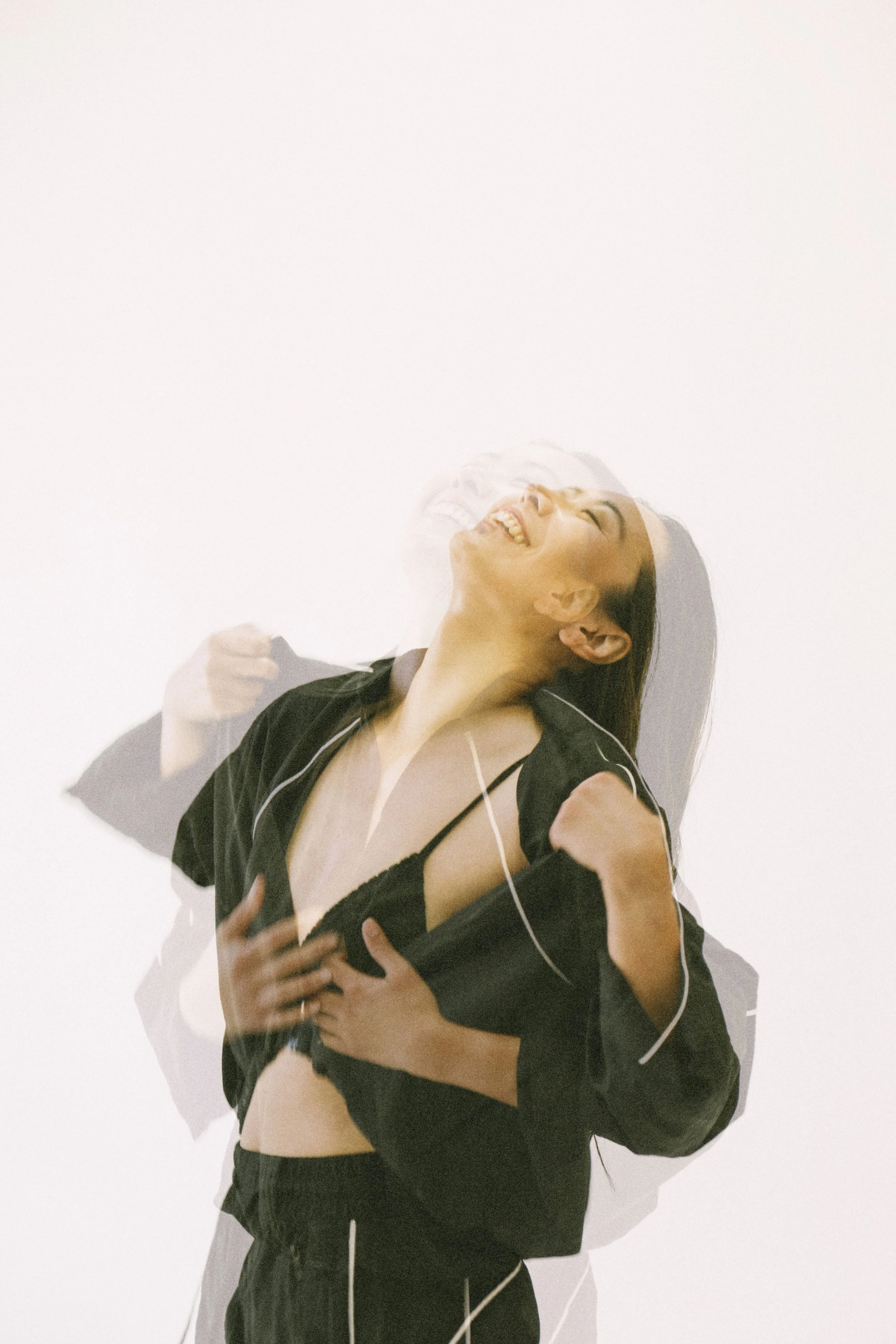 A woman smiling and laughing with her head tilted back, wearing a black top and jacket against a plain white background, with a double exposure effect showing her multiple overlapping images.