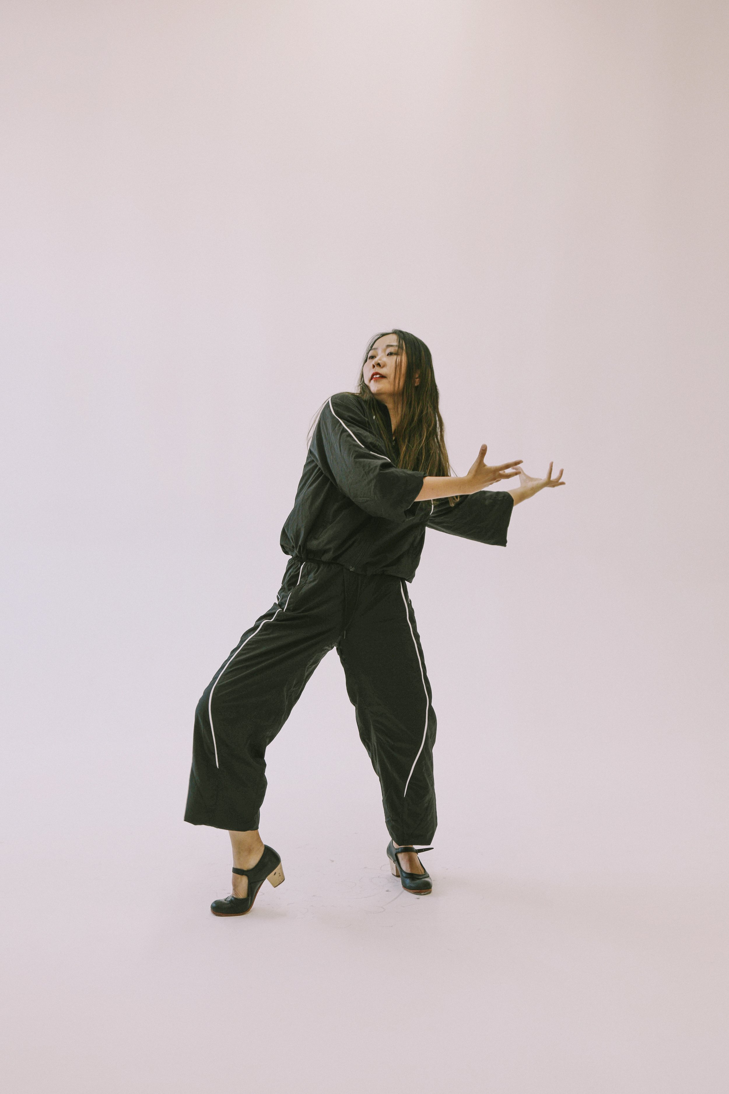 A woman in a black tracksuit and high heels striking a pose against a plain white background.