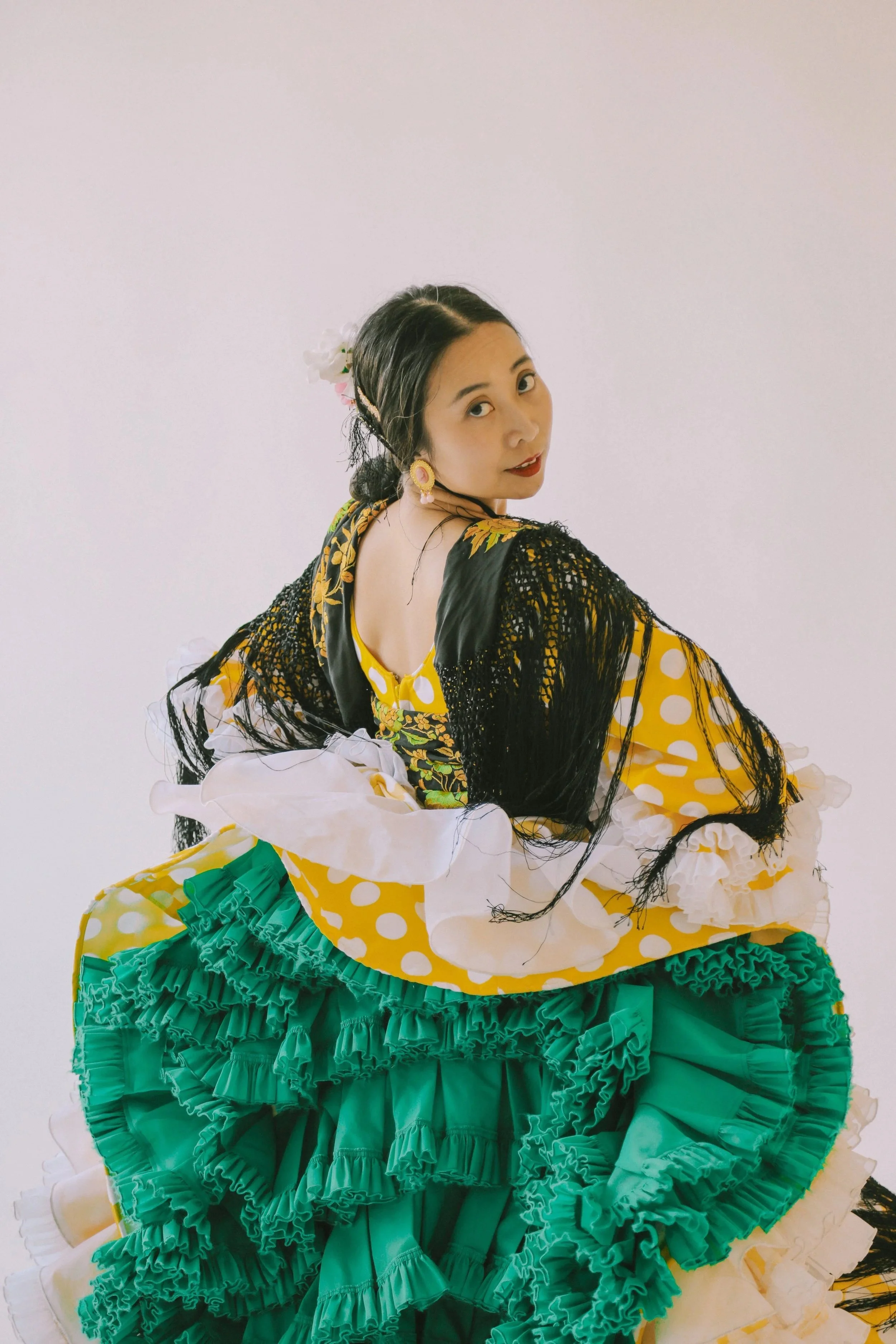 Woman wearing a colorful, traditional dress with bright green ruffled layers, yellow with white polka dots, and a black shawl with fringe, posing against a plain white background.