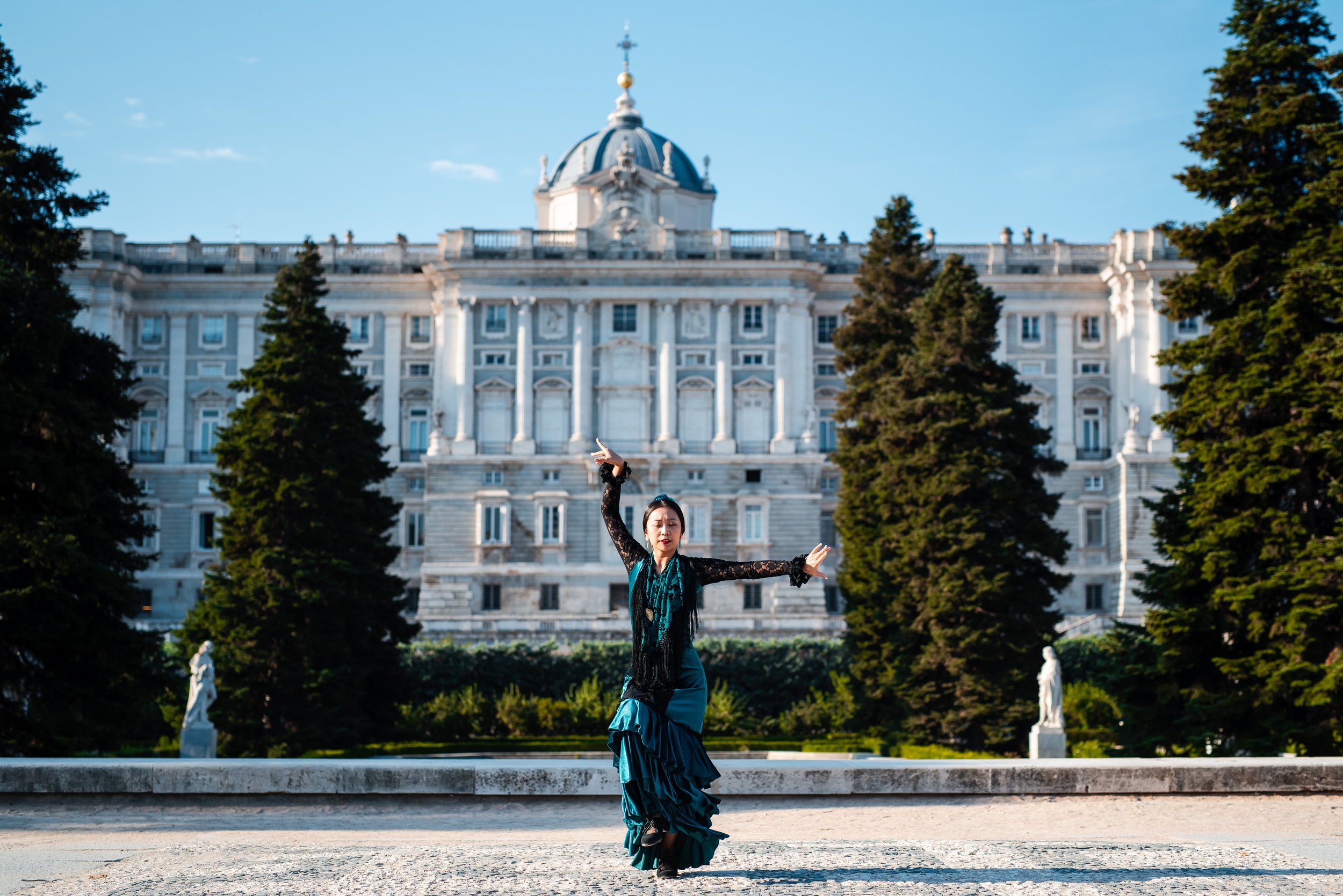 A woman in a black lace top and teal skirt dancing outdoors in front of a large historic building with columns and a dome, surrounded by tall trees.