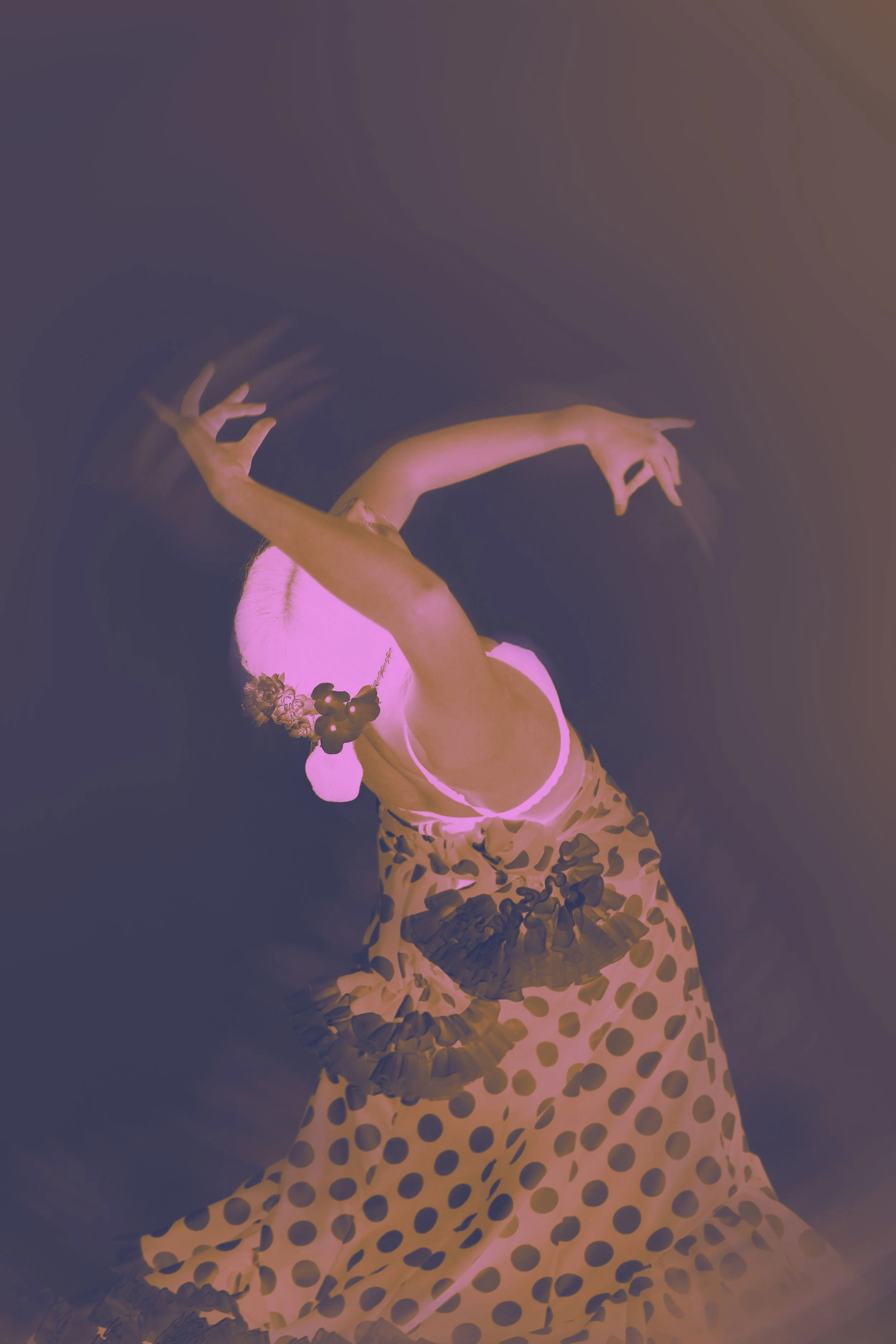 A woman with platinum blonde hair in a bun, wearing a floral headpiece, a pink bra, and a polka dot skirt, is dancing in a dimly lit space. She is positioned with her arm raised above her head and her body gracefully curved, creating an elegant pose.
