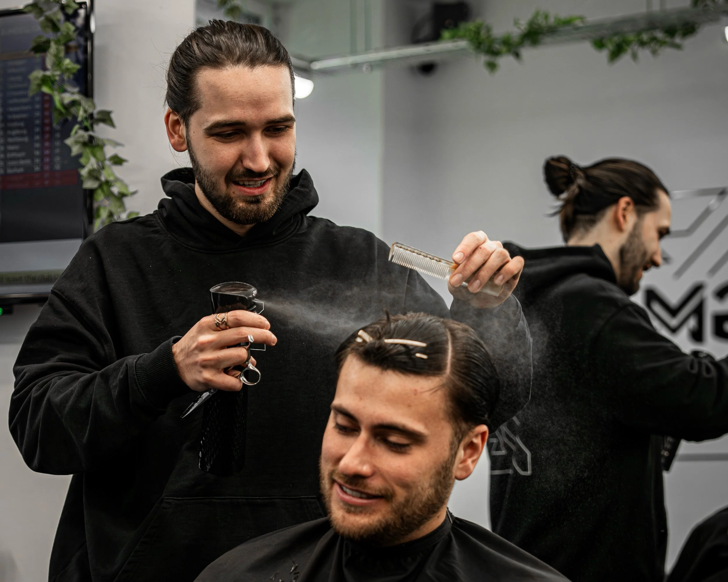 A hairstylist sprays water on a male client's hair while another hairstylist works in the background at a salon.