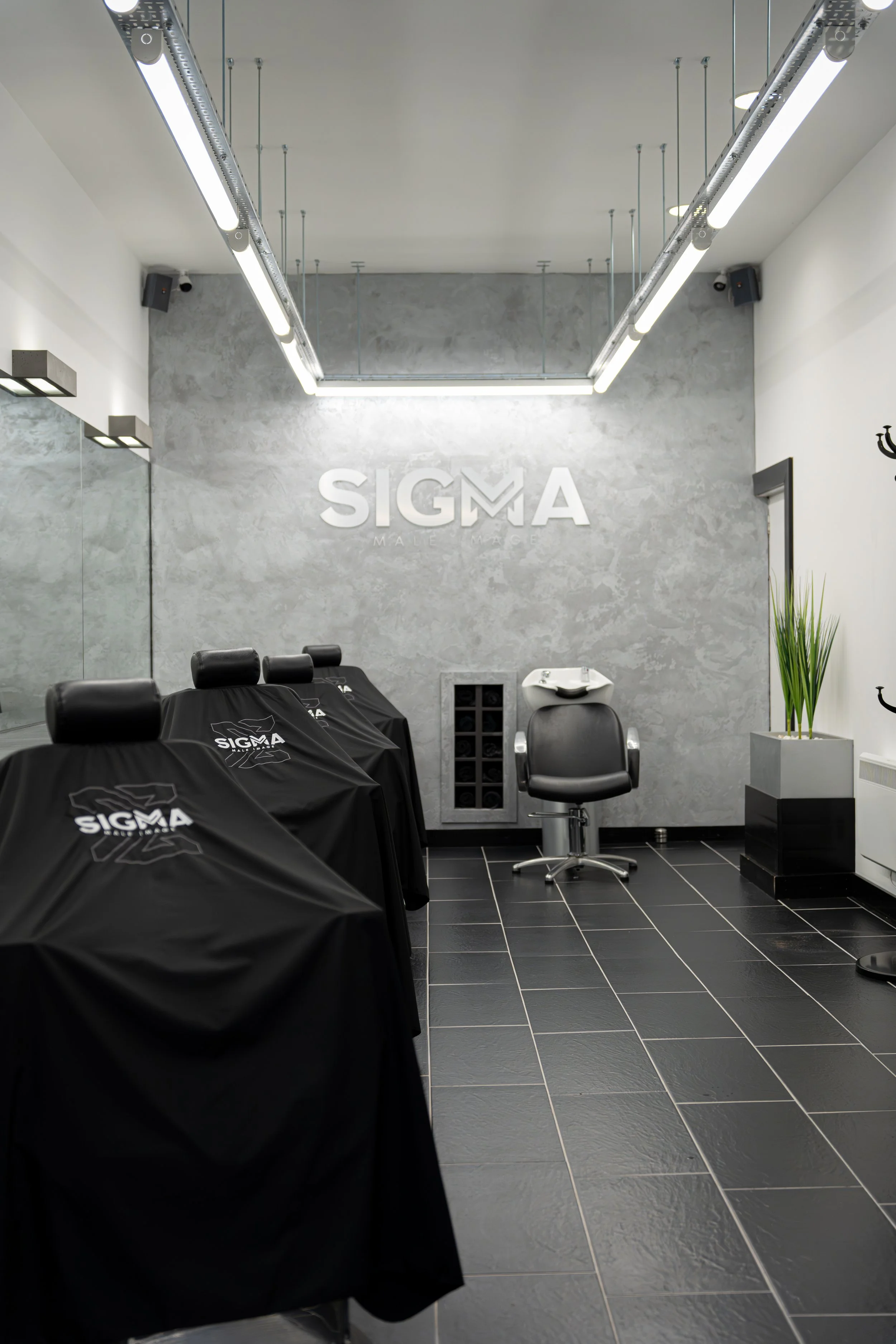 Tattoo studio interior with three black chairs covered in black cloths with the SIGMA logo, a black chair with a sink for tattooing, large mirror, modern lighting, and gray textured wall with SIGMA logo.