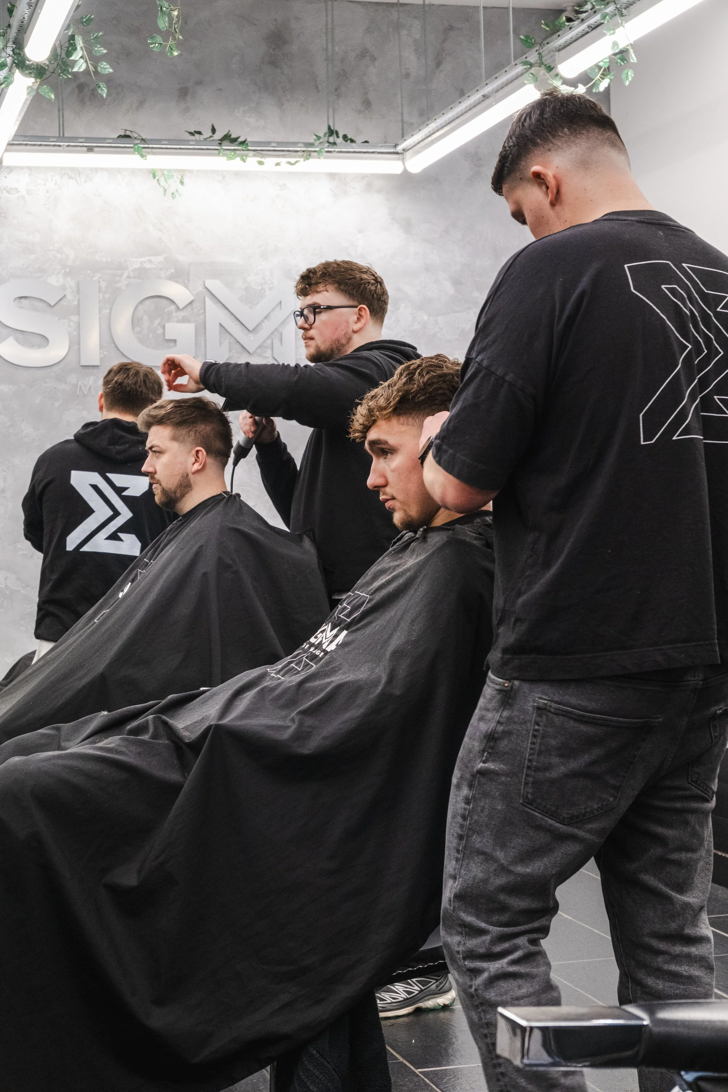 A man receives a haircut at a barbershop with three other men in the background, all wearing black hoodies with a stylized logo.