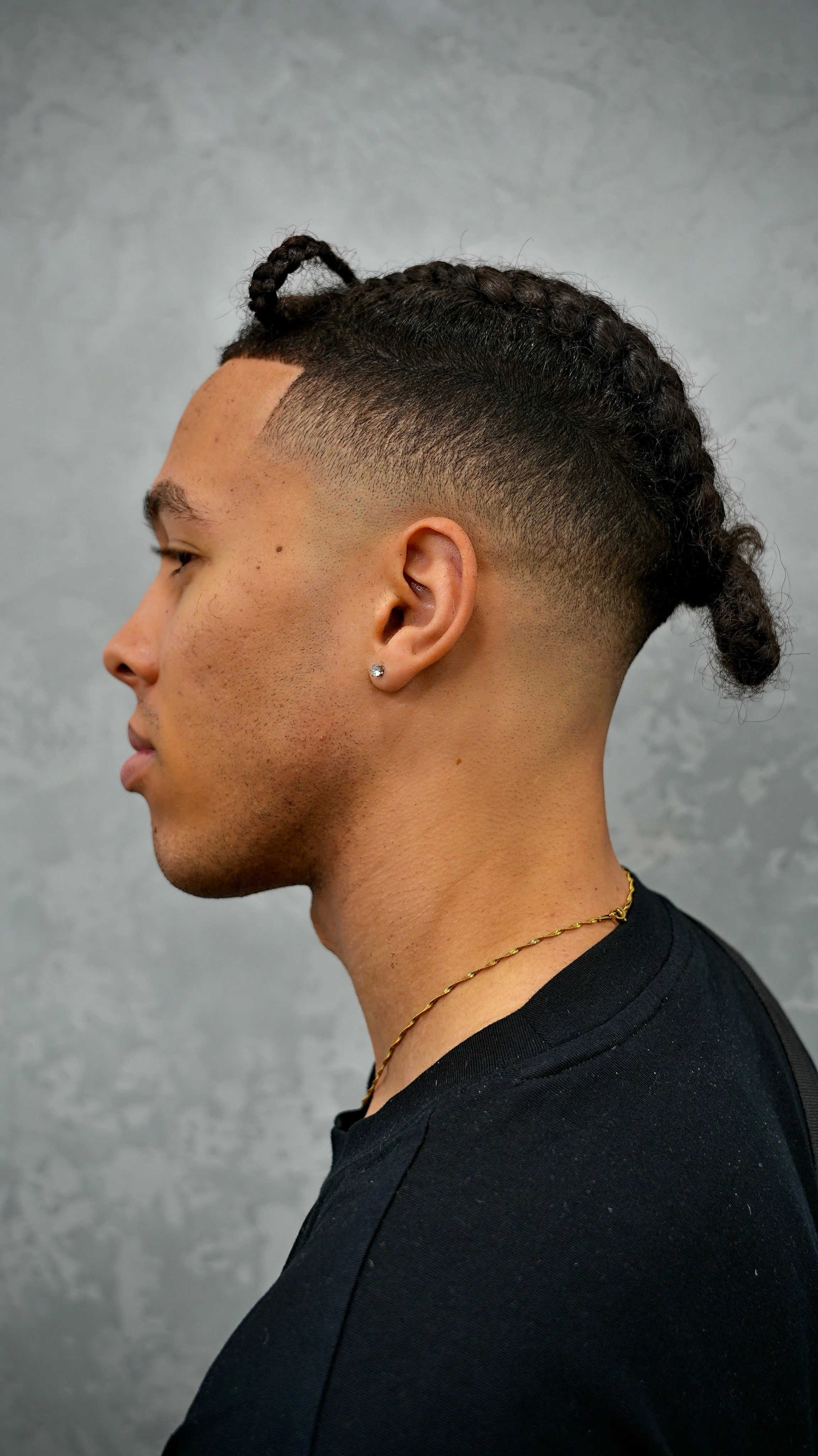 Side profile of a man with a fade haircut and braided hair on top, wearing a black shirt, earrings, and a gold necklace, against a gray background.