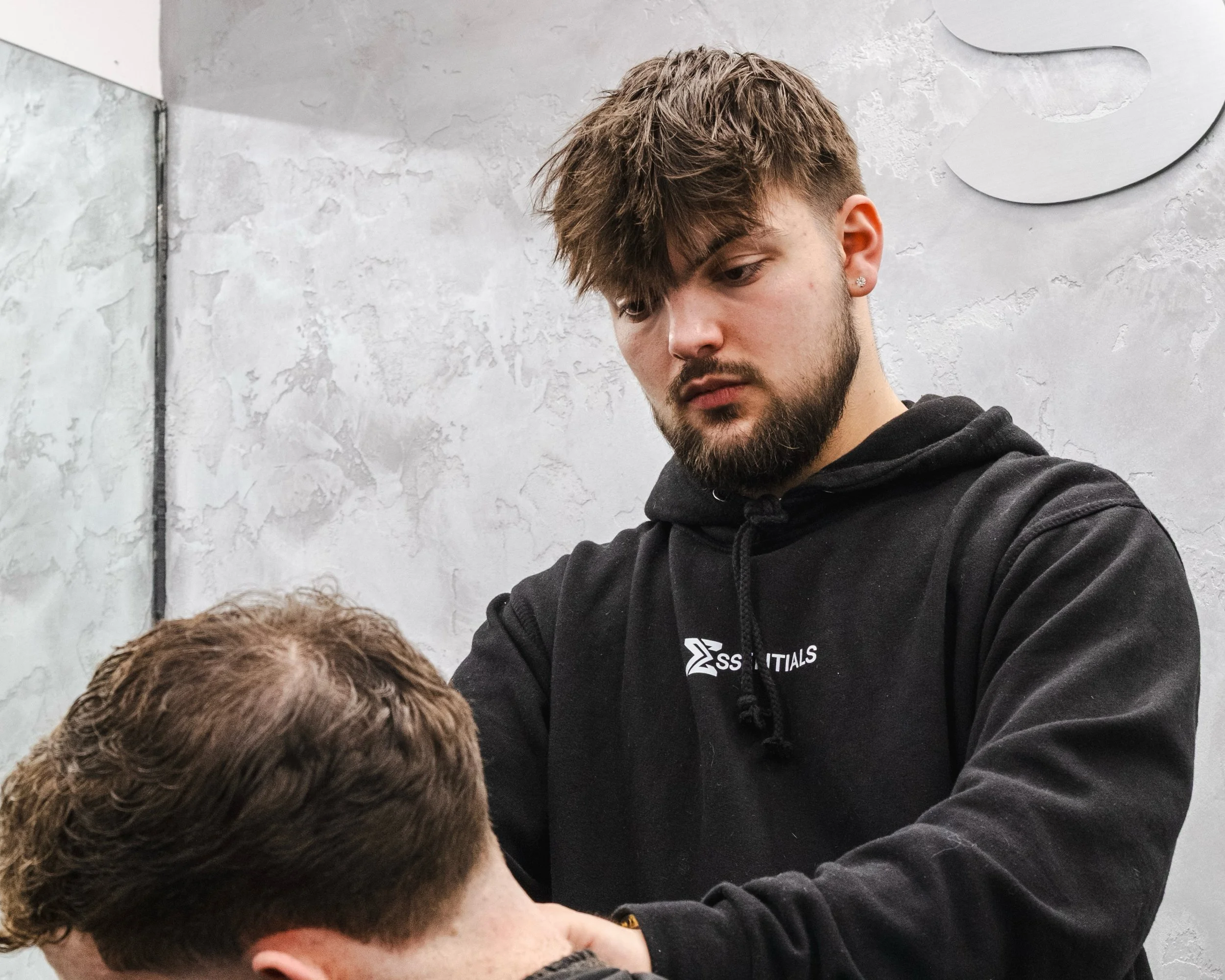 A young man with a beard and brown hair, wearing a black hoodie, appears to be practicing massage or physical therapy on another person with short brown hair, whose head is reclined.