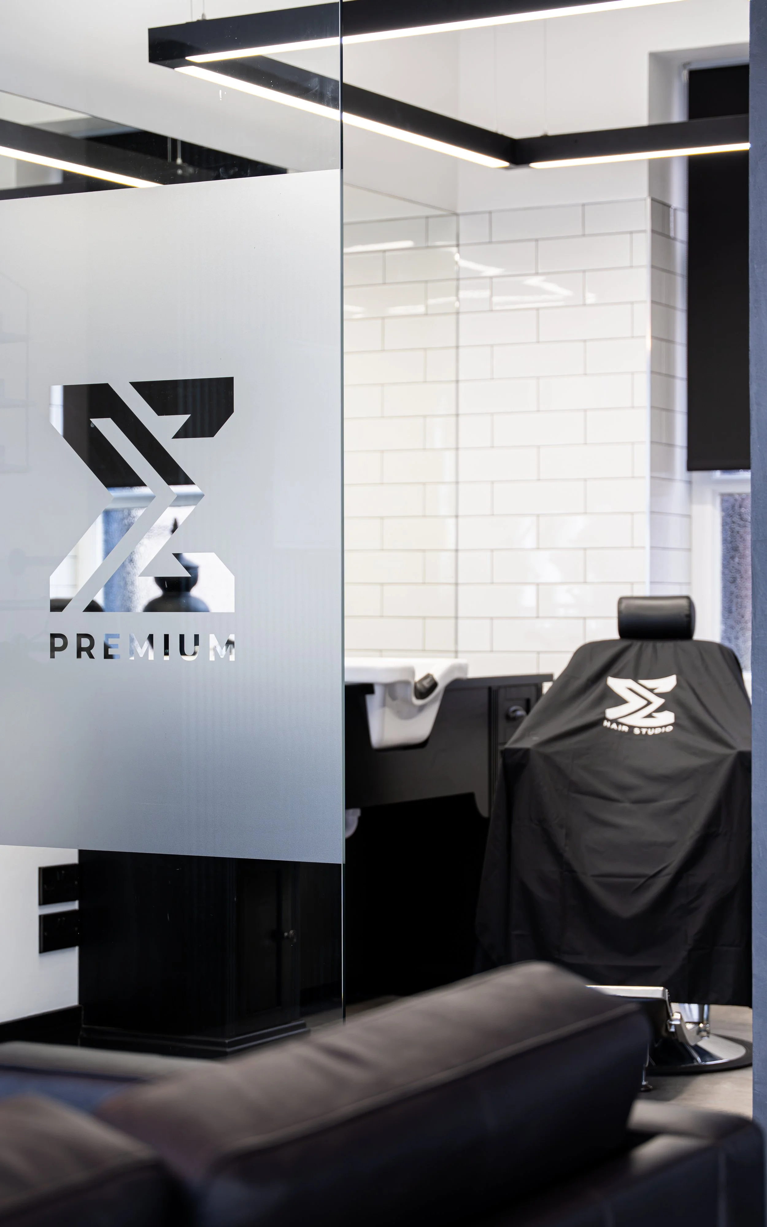 View inside a modern barber shop with a salon chair, a mirror, and a glass partition that has the word "PREMIUM" and a stylized logo on it. The space has white tiled walls and black accents.