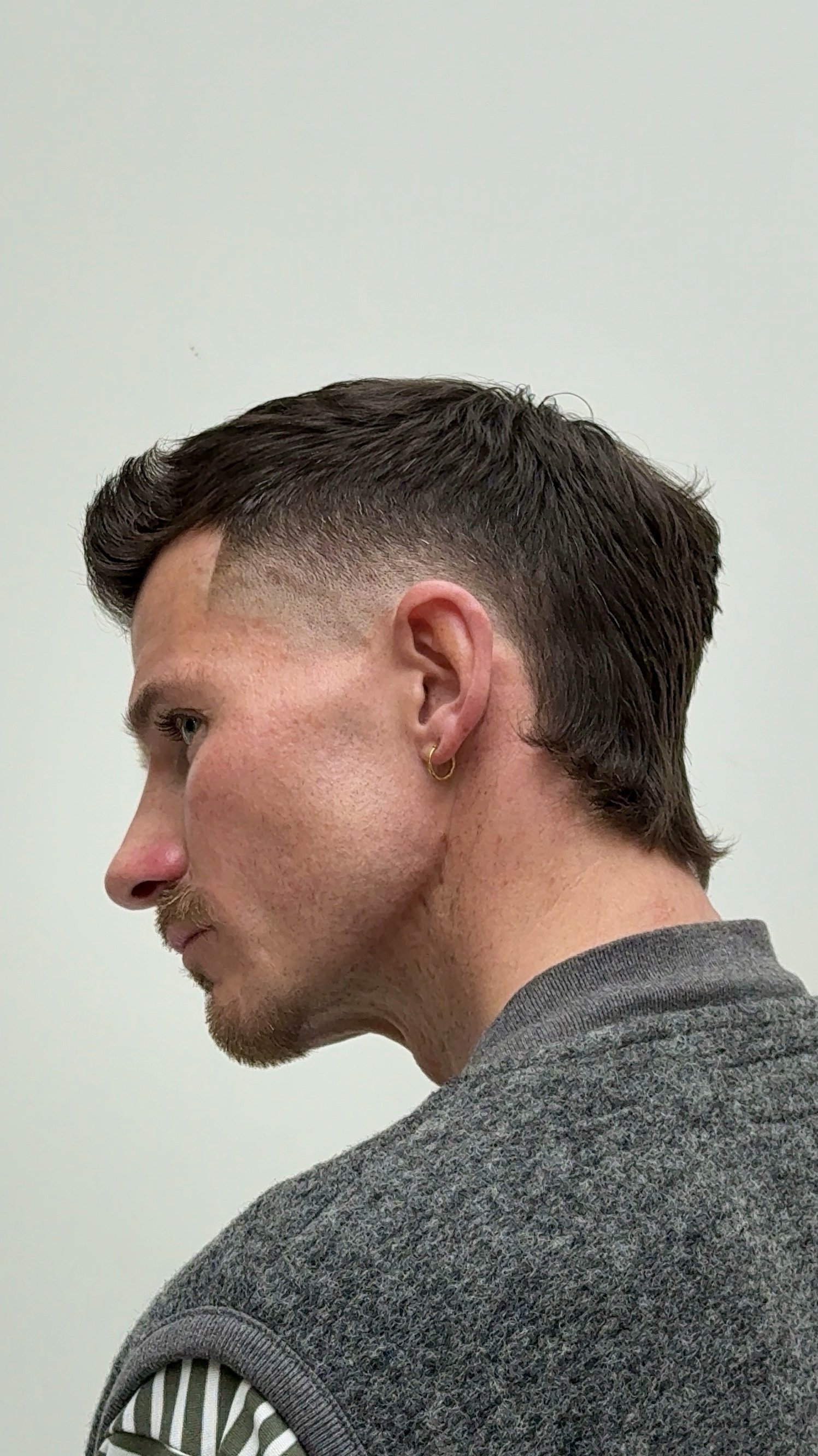 Side profile of a man with dark, short, and styled hair, wearing a gray sweater with a striped sleeve and a gold hoop earring in his ear, against a plain light background.