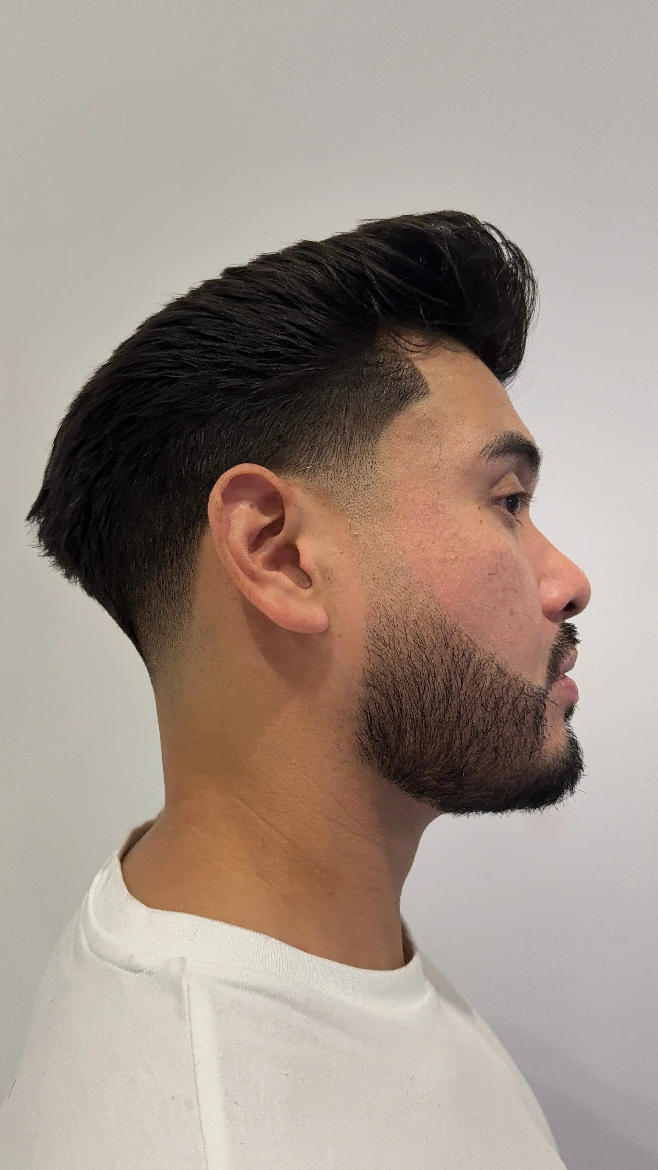 Profile of a man with a dark, slicked-back hairstyle, fade haircut, and beard, wearing a white shirt, against a plain light-colored background.