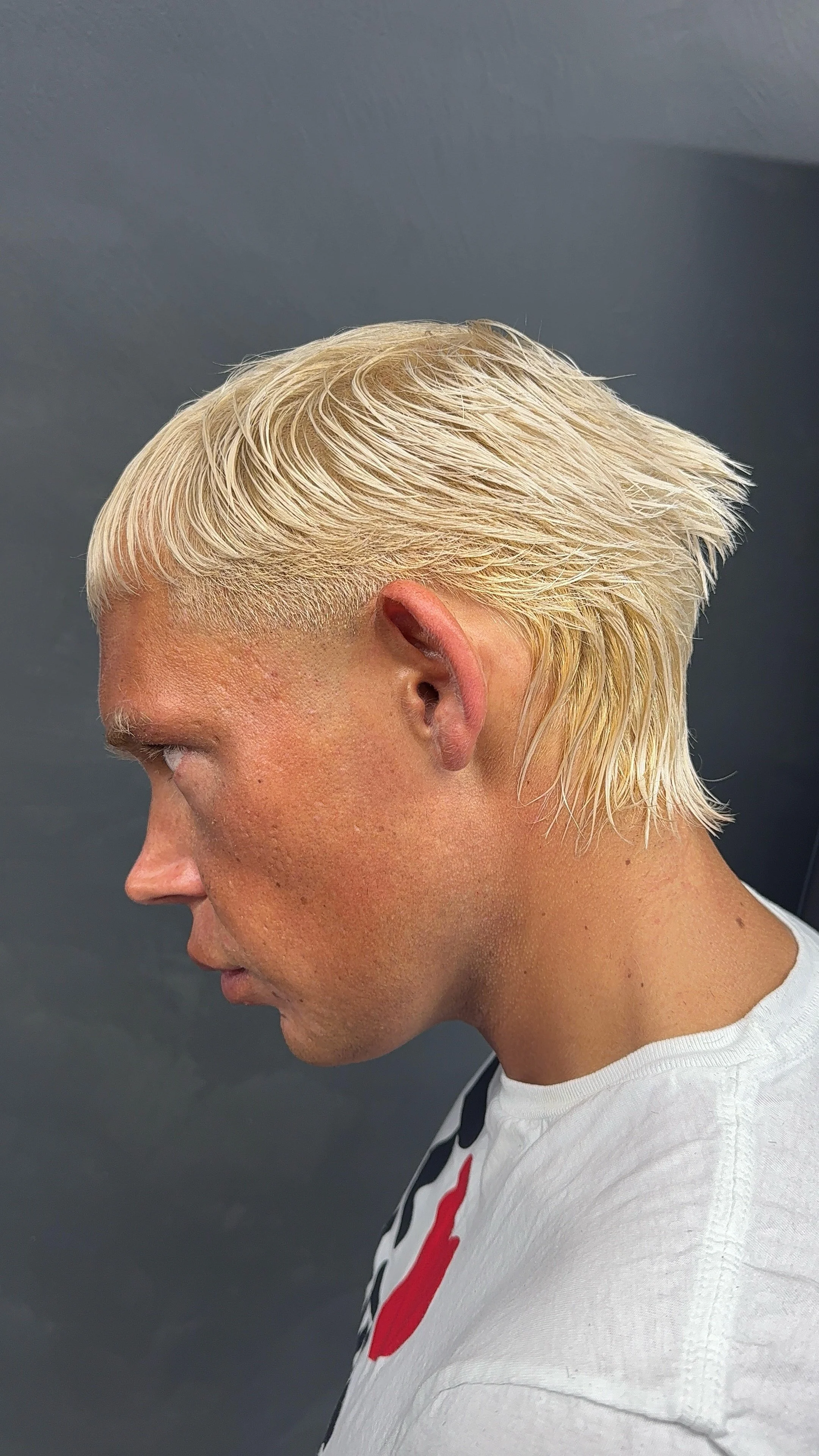 Side view of a person with short, platinum blonde hair and light skin, wearing a white shirt with a graphic design, against a dark background.