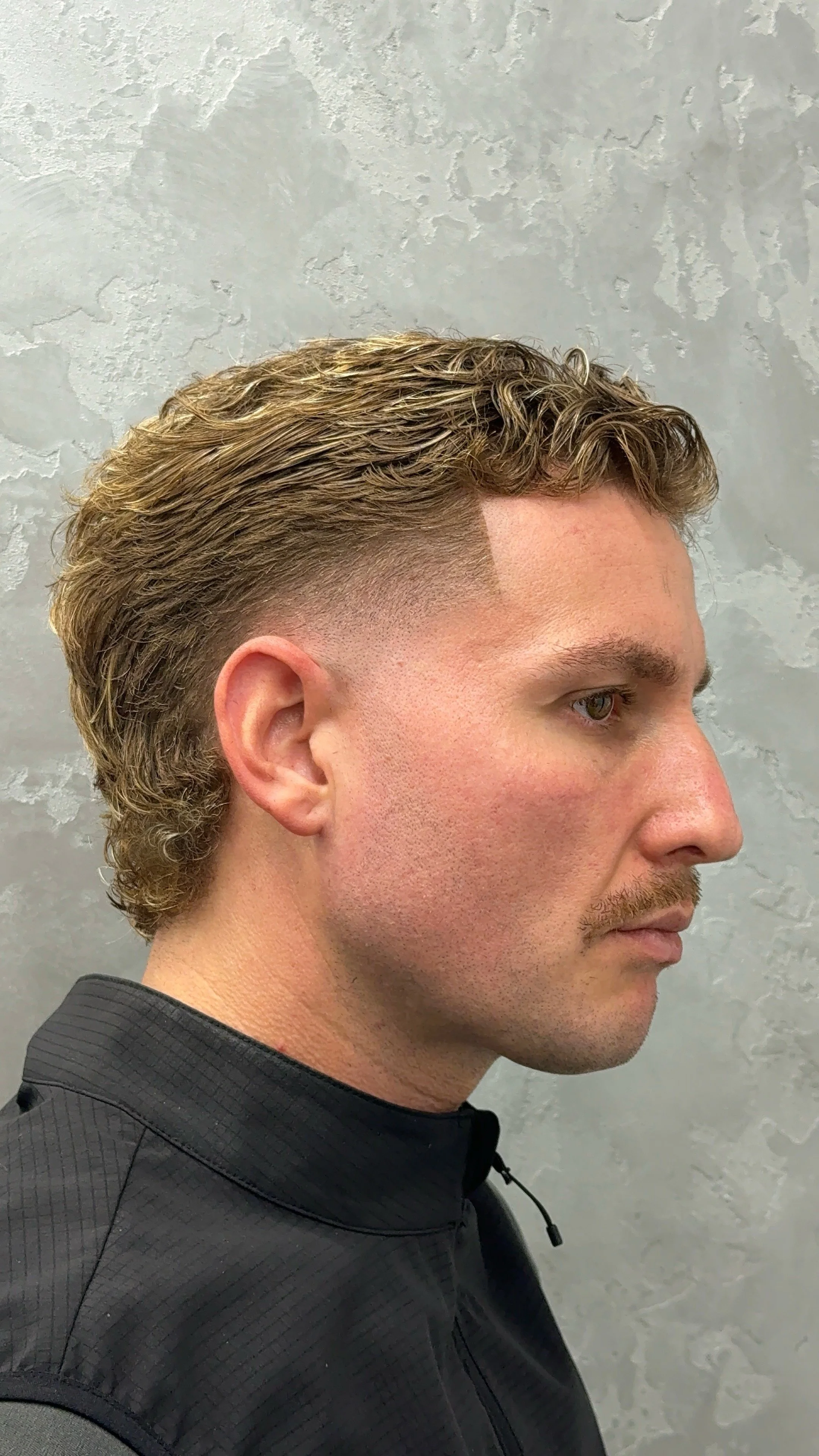 Side view of a man with styled, curly hair and a fade haircut, wearing a black zip-up jacket against a textured gray wall.