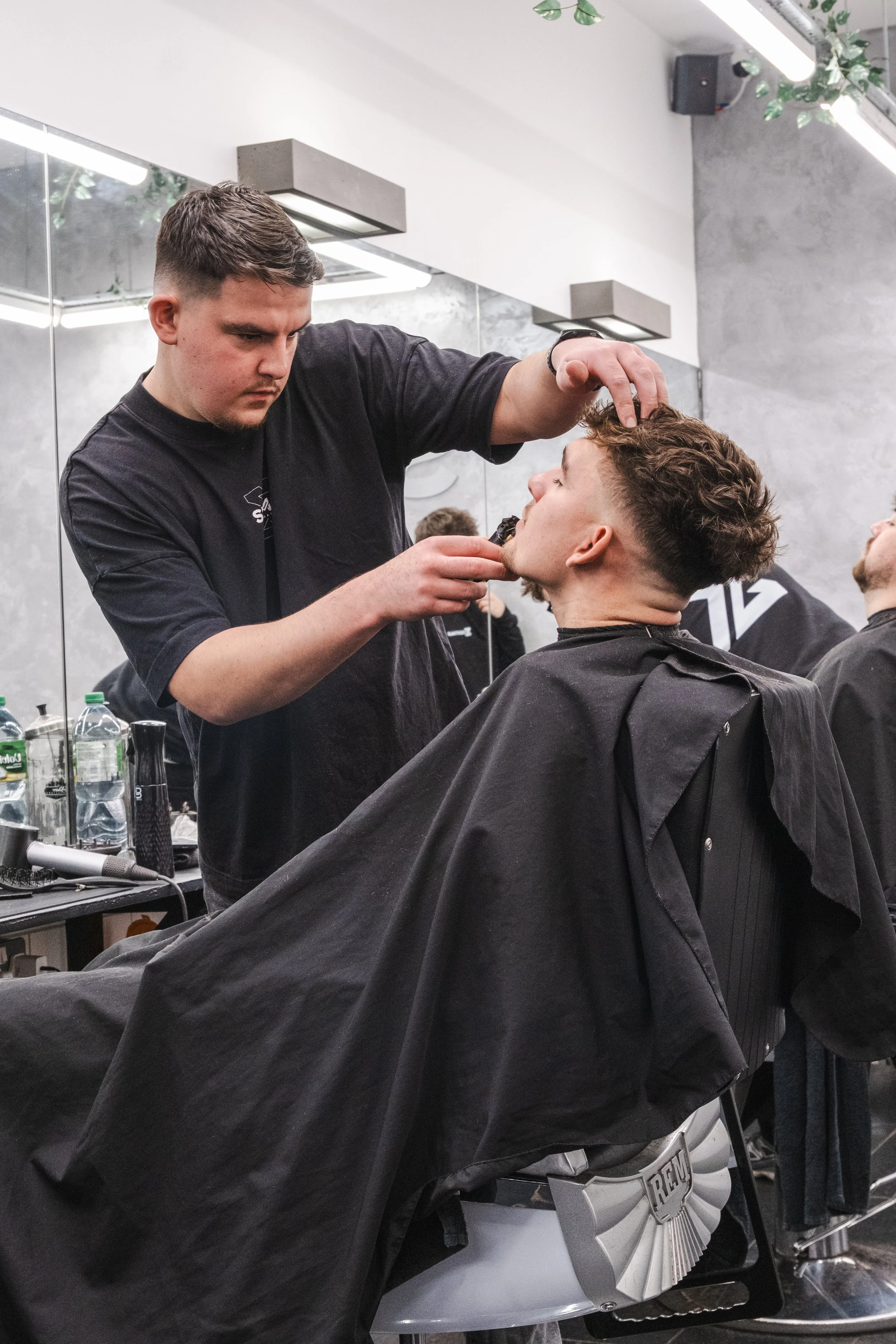 Barber cutting a young man's hair in a salon with mirrors and bottles visible on the counter.