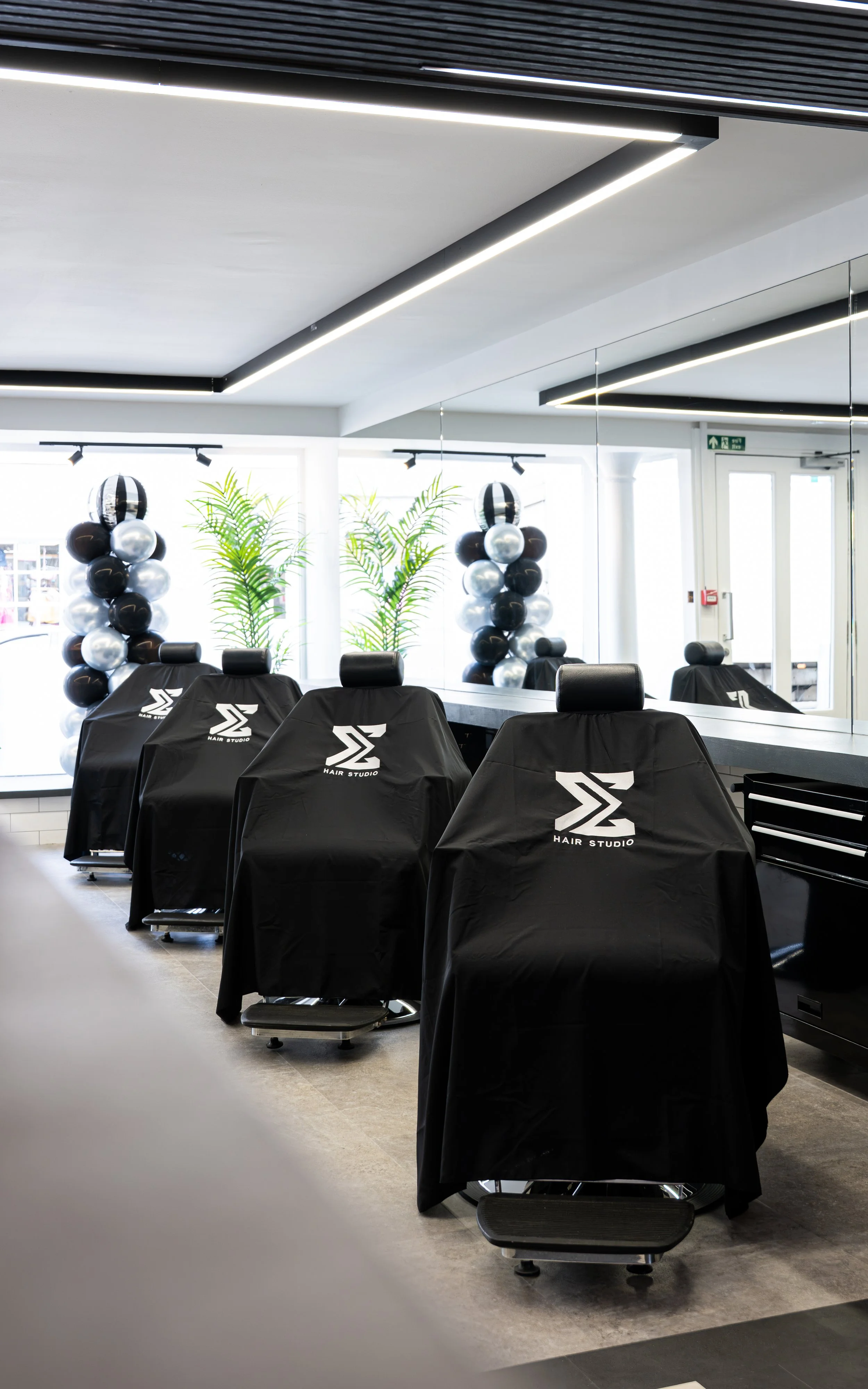 Empty barbershop with black chairs covered with black capes featuring 'Hair Studio' logo, large windows with plants outside, balloons in black, silver, and white, and modern ceiling lighting.