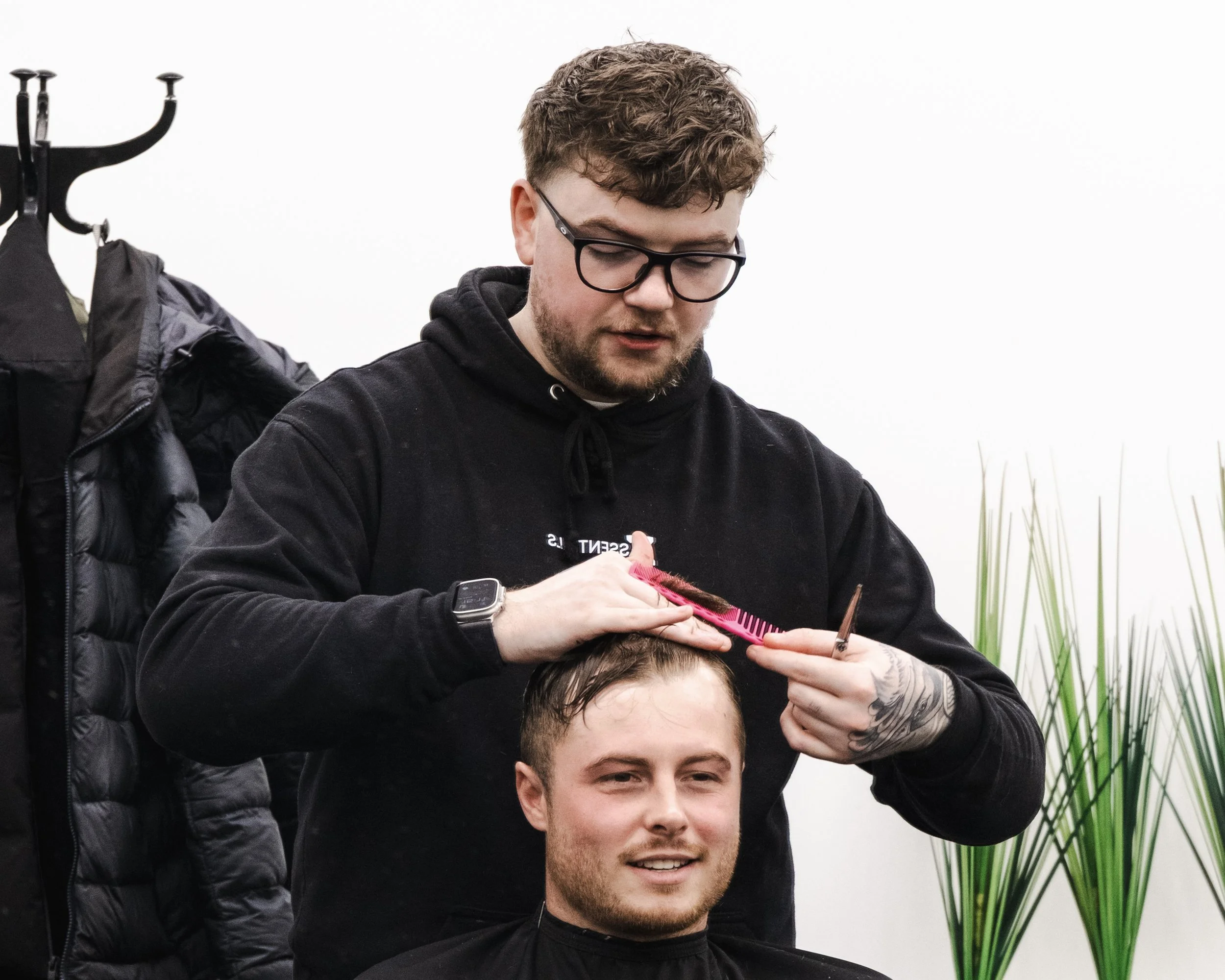 A barber cutting a man's hair in a salon with white walls and a potted plant in the background.