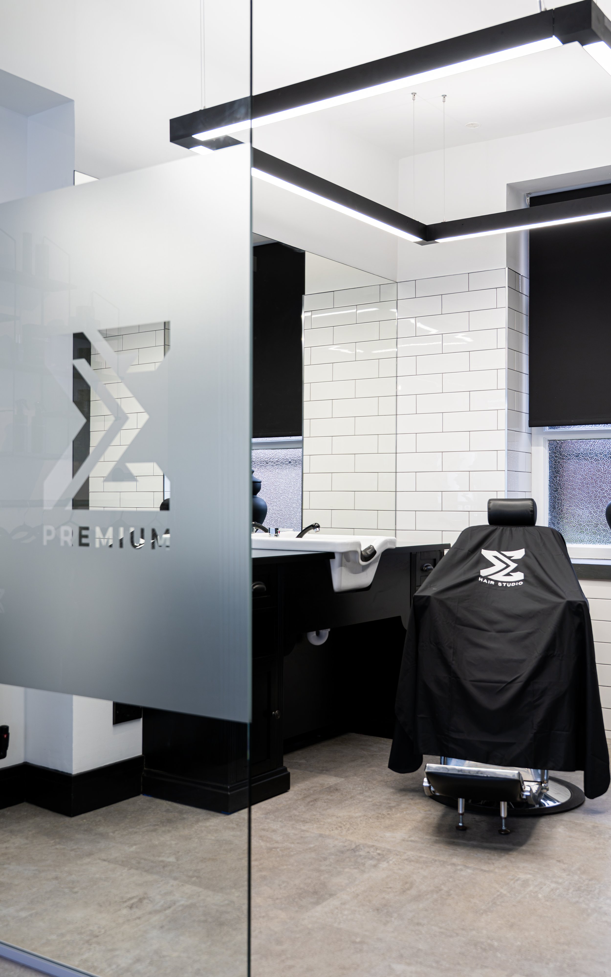 A modern barber shop with white tiled walls, a black barber chair with a black cover that has white logo, a white countertop, and a large window with sheer curtain. Modern square hanging light fixture overhead.