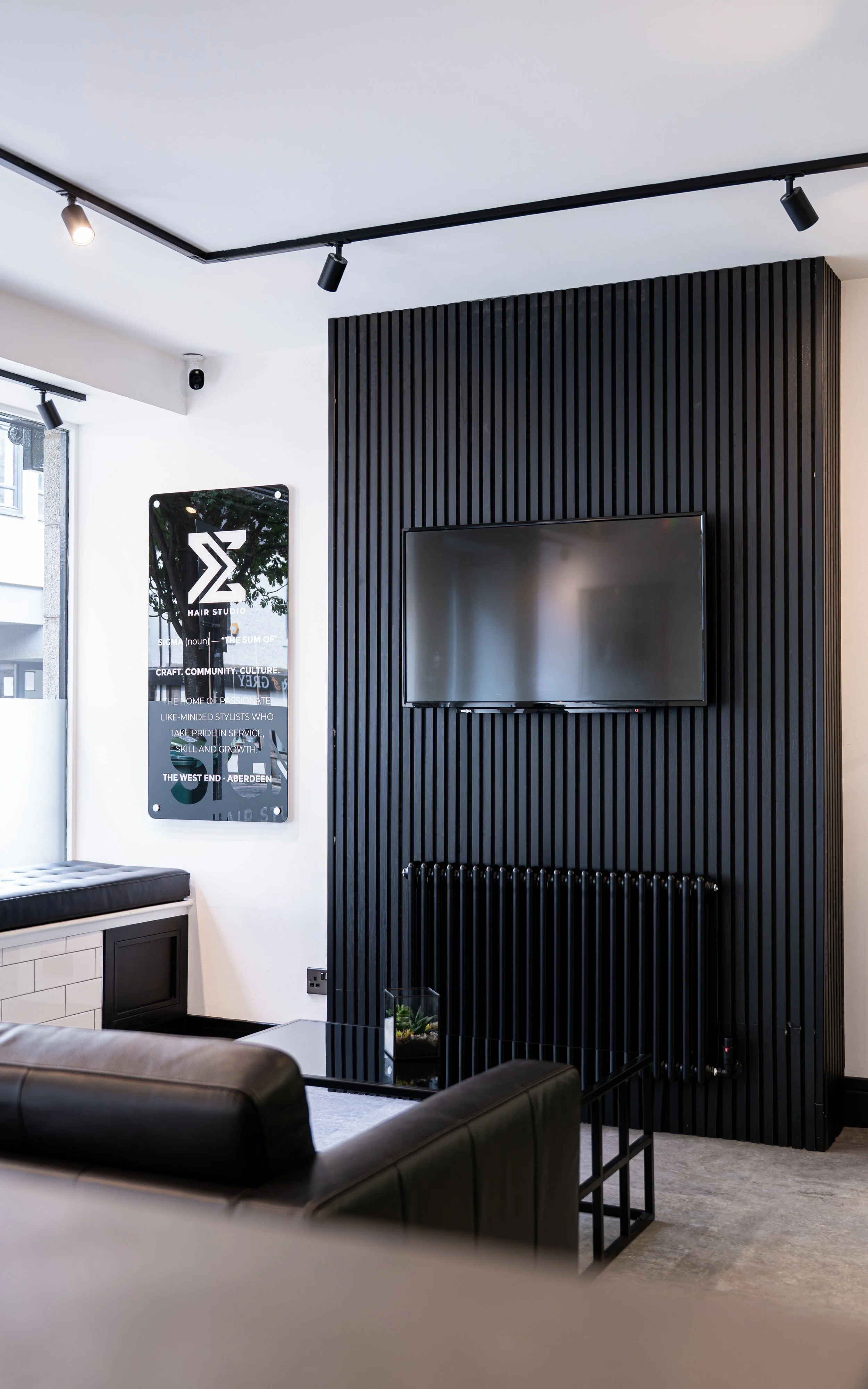 Modern interior with a black slatted wall, a mounted flat-screen TV, a black radiator, a small glass terrarium, and part of a leather sofa.