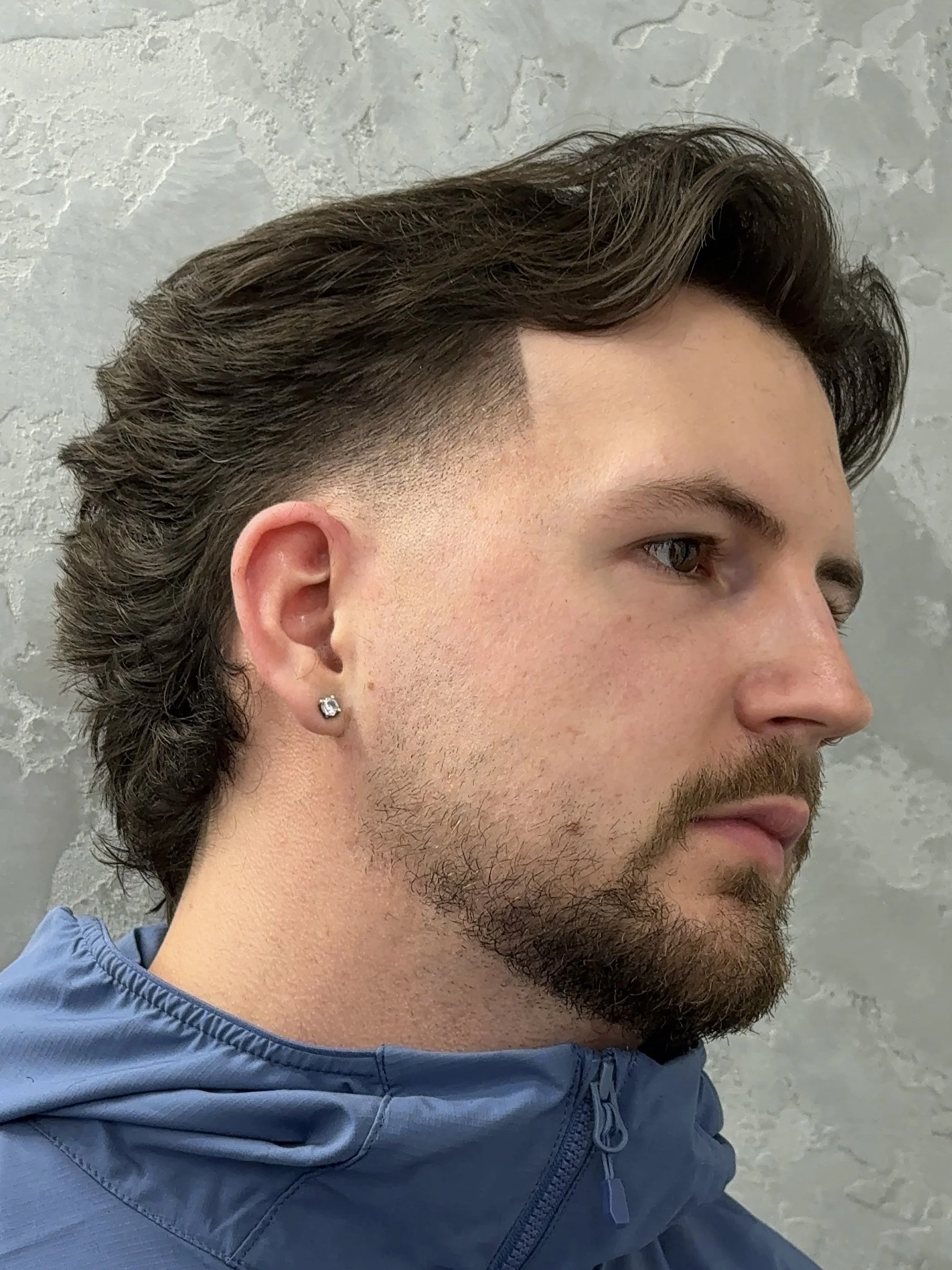 A young man with a full beard and mustache, wearing a small earring, seen in profile. He has styled brown hair with a faded side and is wearing a light blue jacket. The background appears to be a gray textured wall.