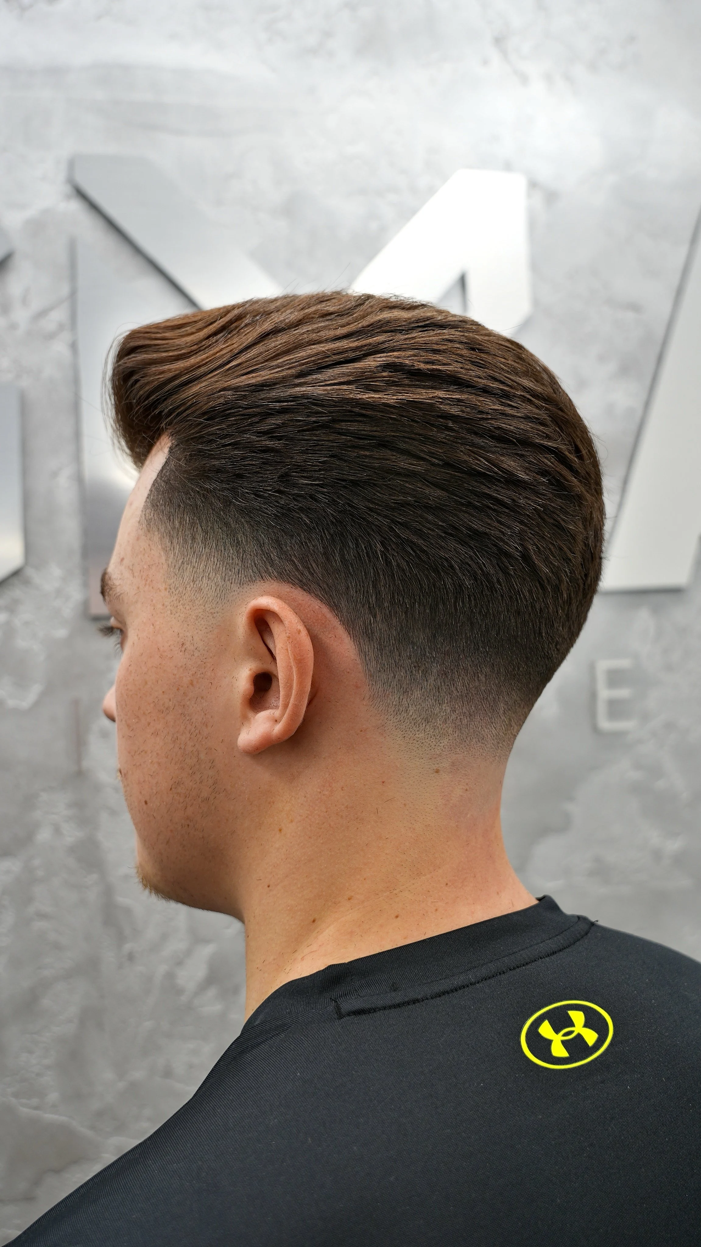 Side view of a young man's hairstyle with a fade, dark brown hair, and a textured top, wearing a black Under Armour shirt.