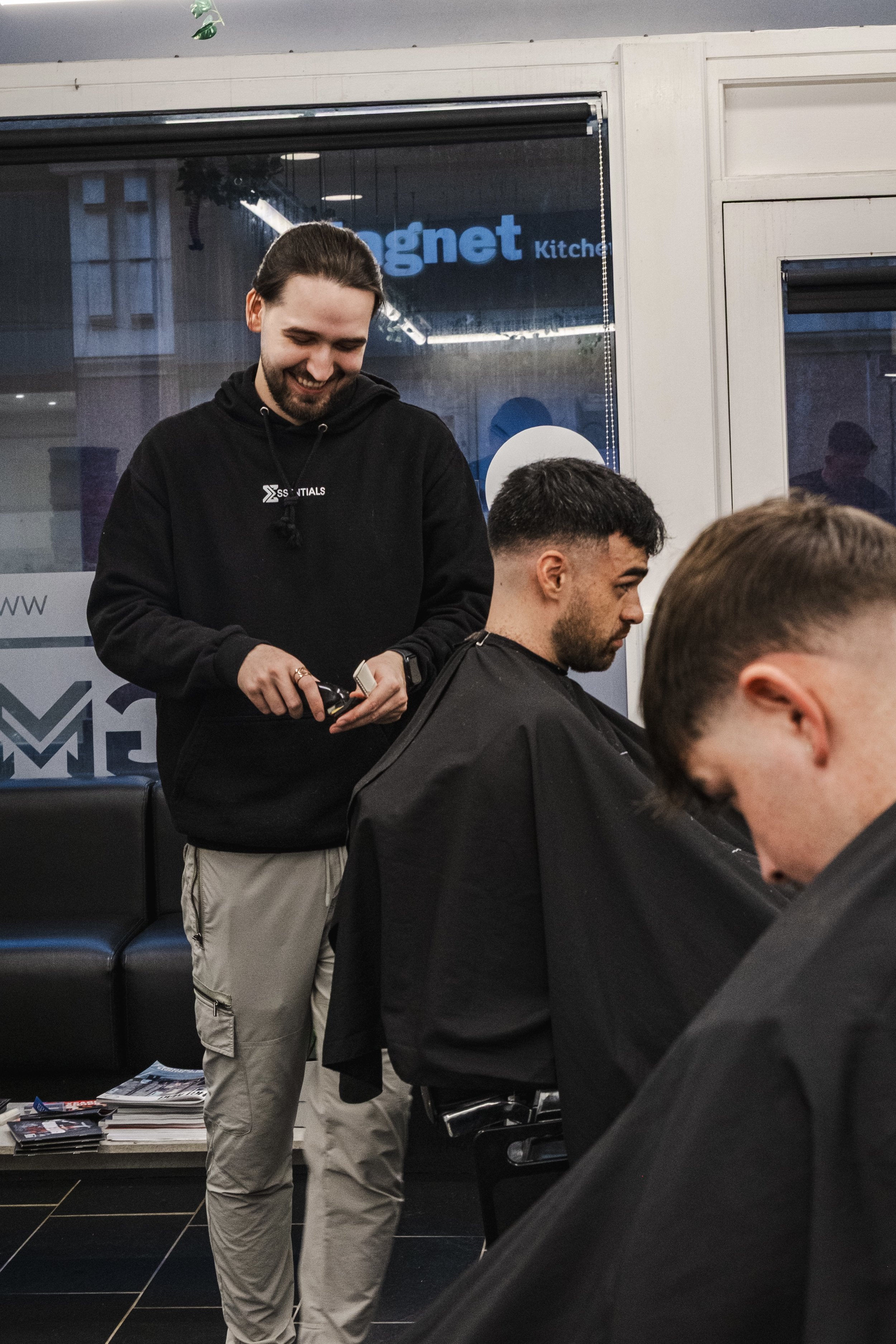 A barber giving a haircut to a male customer in a barbershop with other clients nearby.
