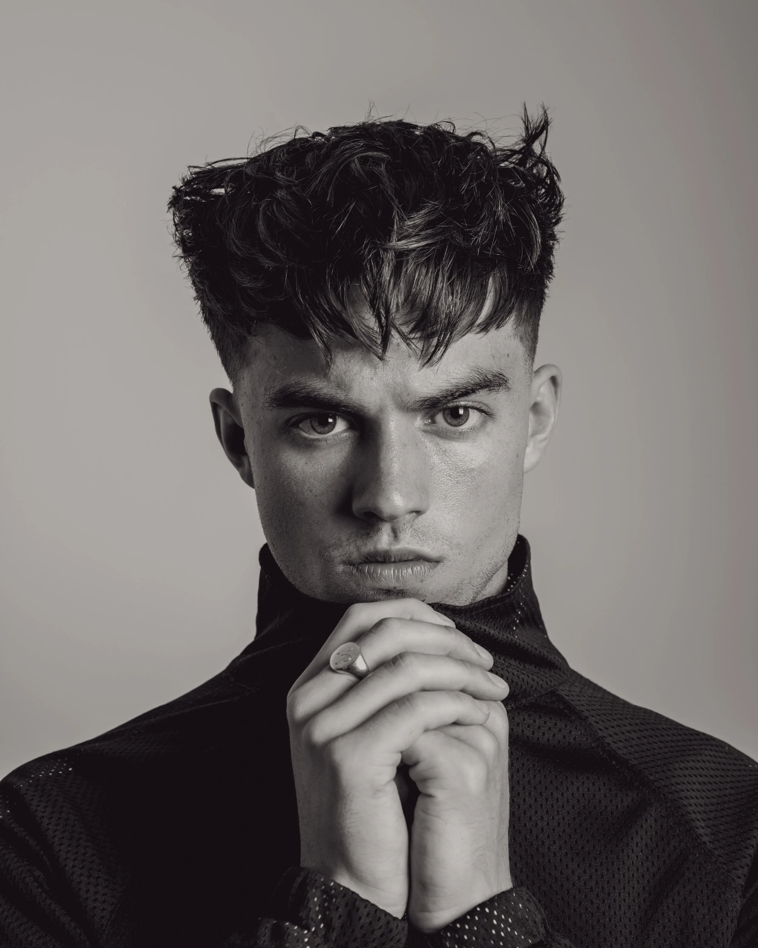 Black and white portrait of a young man with styled hair, intense gaze, and hands clasped in front of his chin, wearing a patterned jacket.