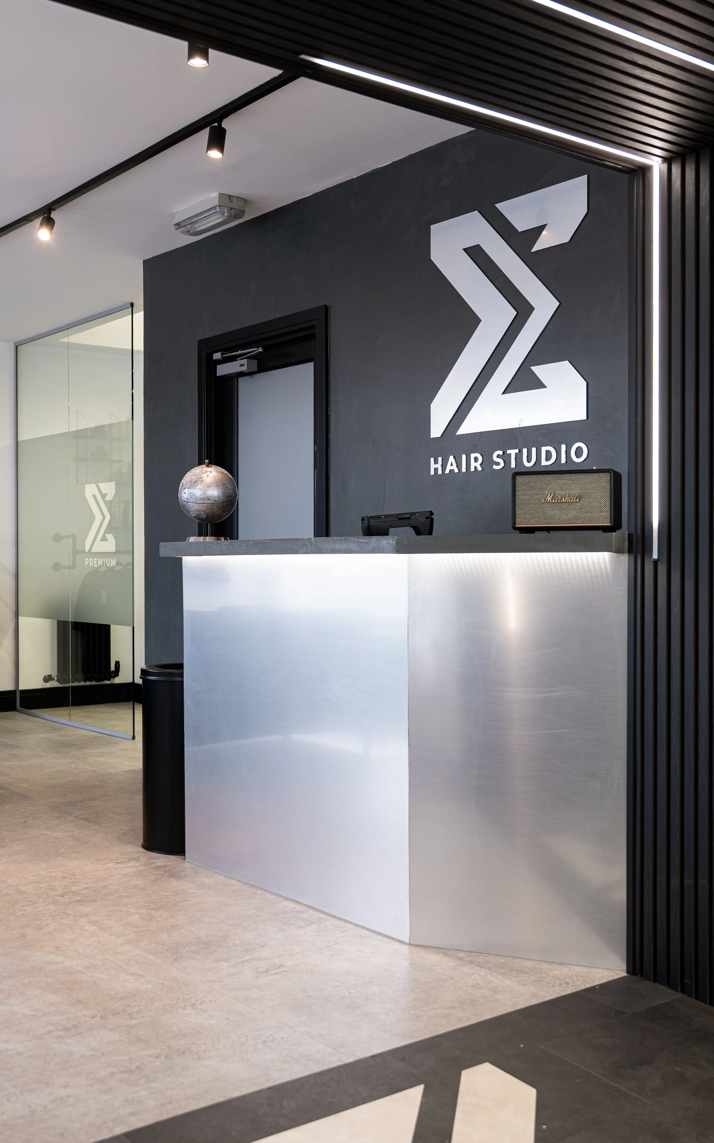 Modern reception area of a hair studio with a metallic counter, a black wall with the 'Z2 Hair Studio' logo in white, a globe sculpture, and an amplifier on the counter. There is a glass door with a similar logo on it.