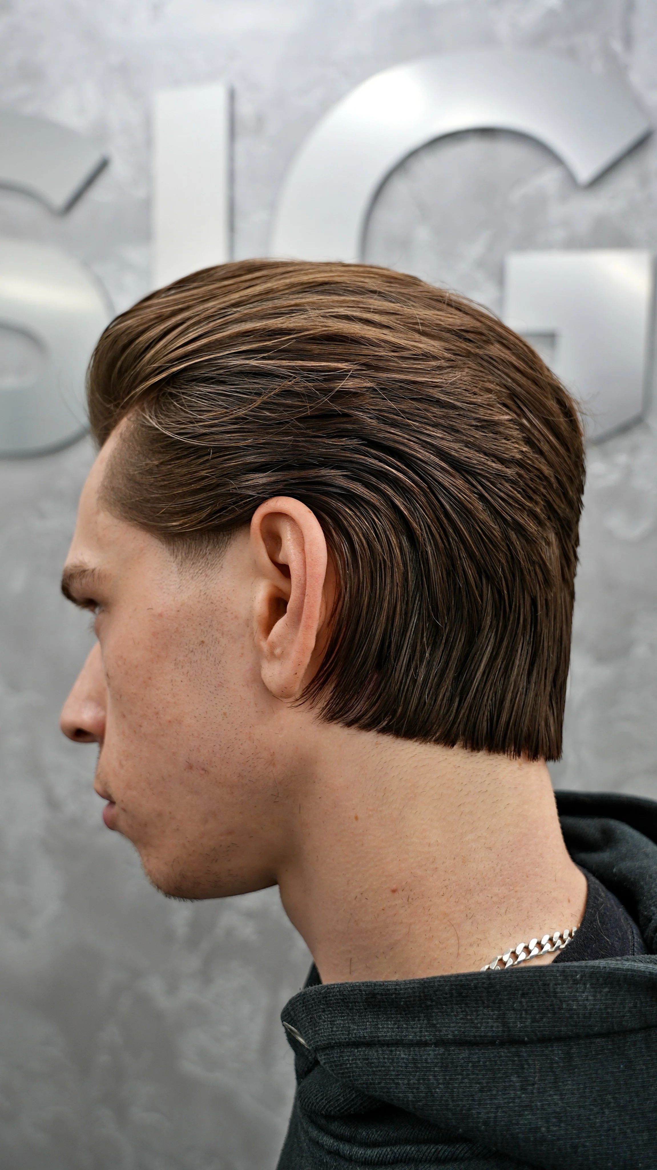 Side view of a young man with a sleek, styled brown haircut, wearing a black hoodie and a silver chain, standing in front of a gray wall with metallic letters.