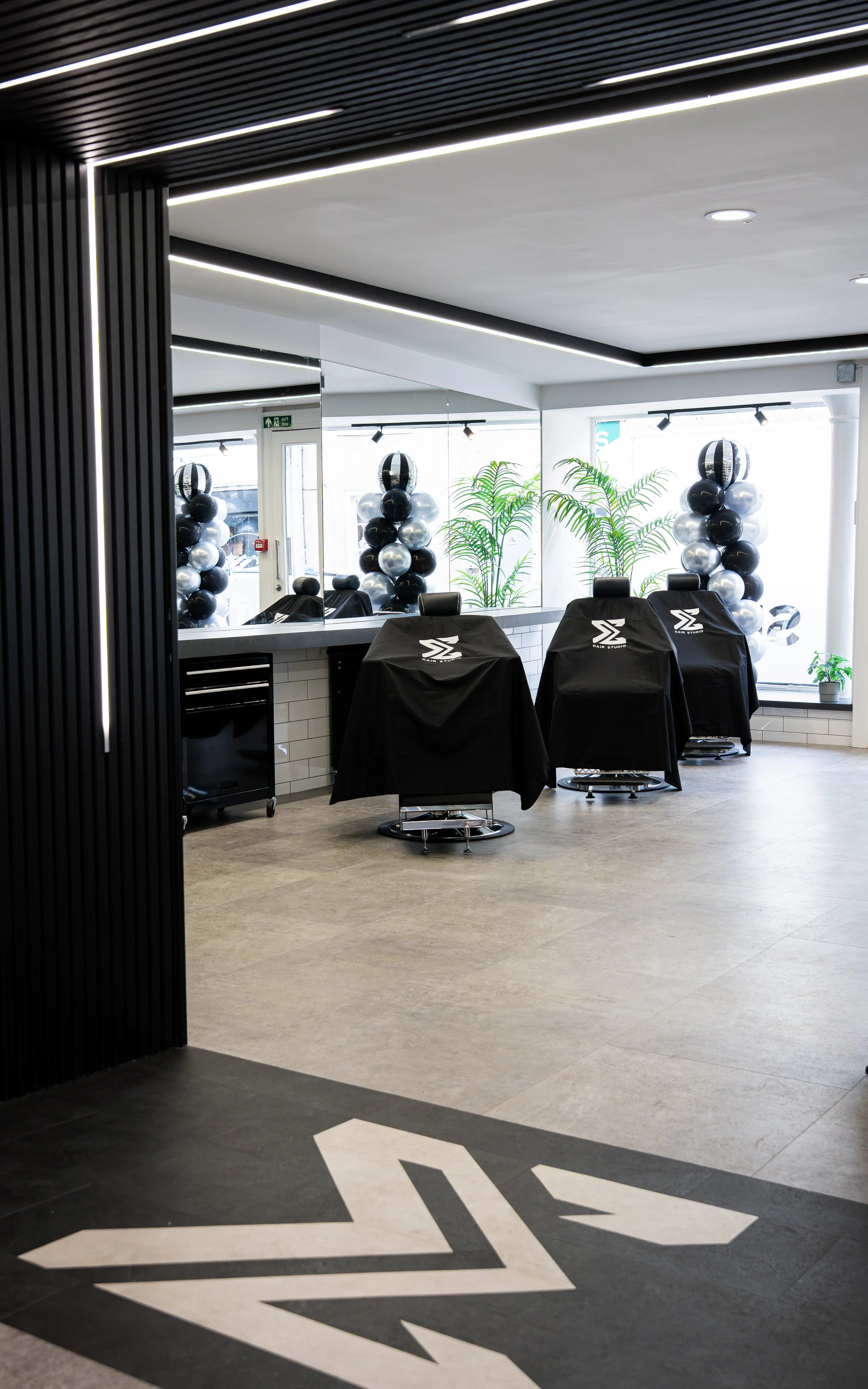 Modern salon with black and white decor, black barber chairs covered with black capes featuring a white logo, large mirror with round decorations made of black, silver, and white balloons, and tall green plants in the background.